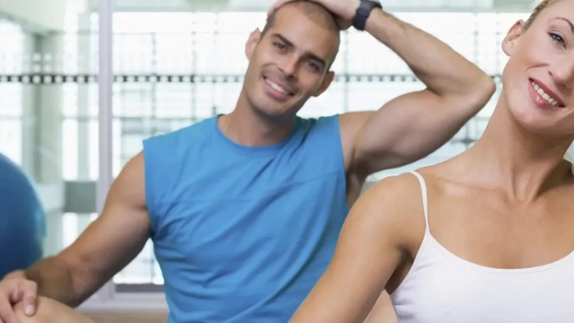 Smiling man and woman stretching neck muscles in a bright gym with exercise ball in background.
