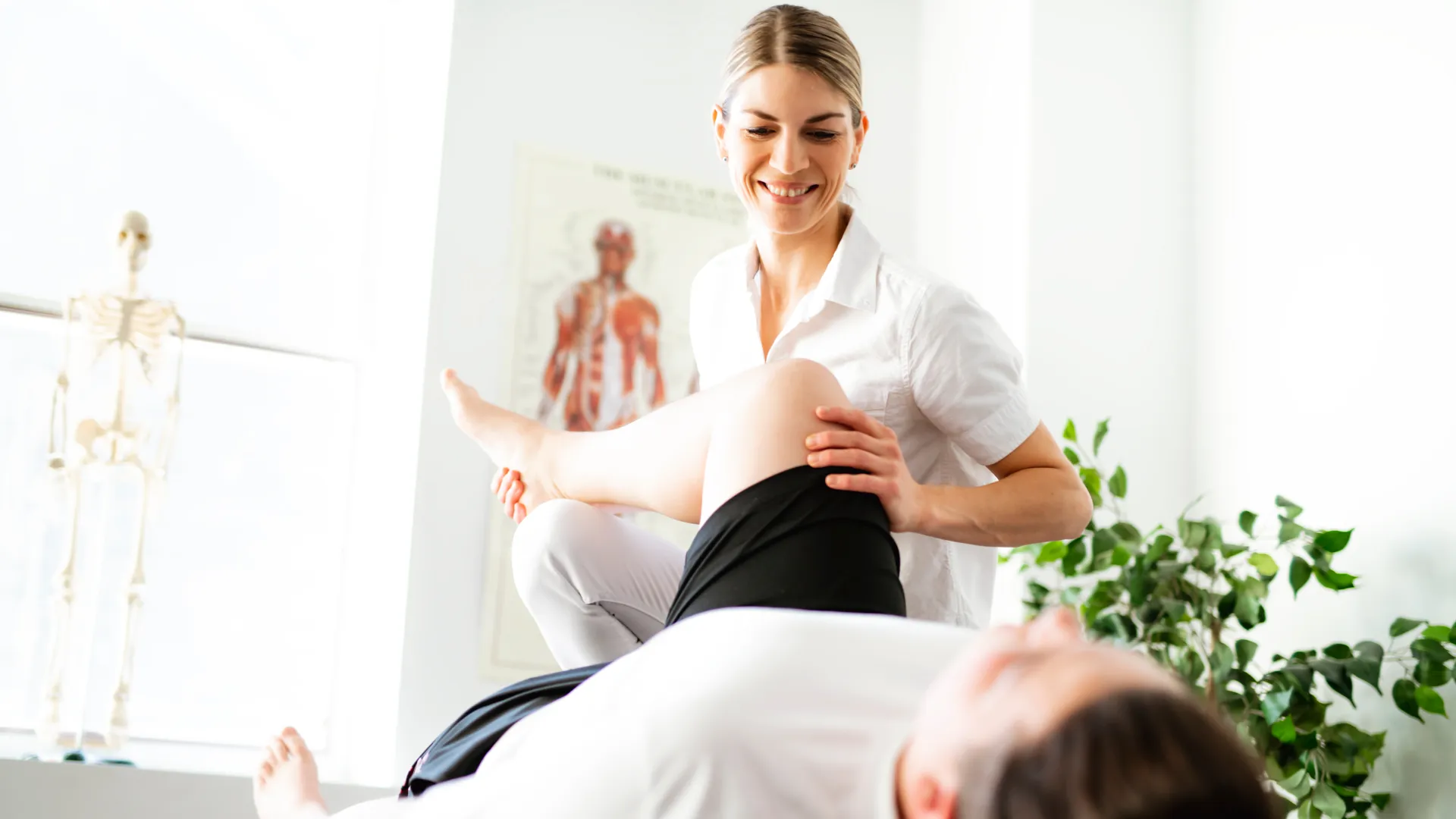 Physiotherapist smiling and stretching patient's leg in a bright clinic with anatomical models and plants