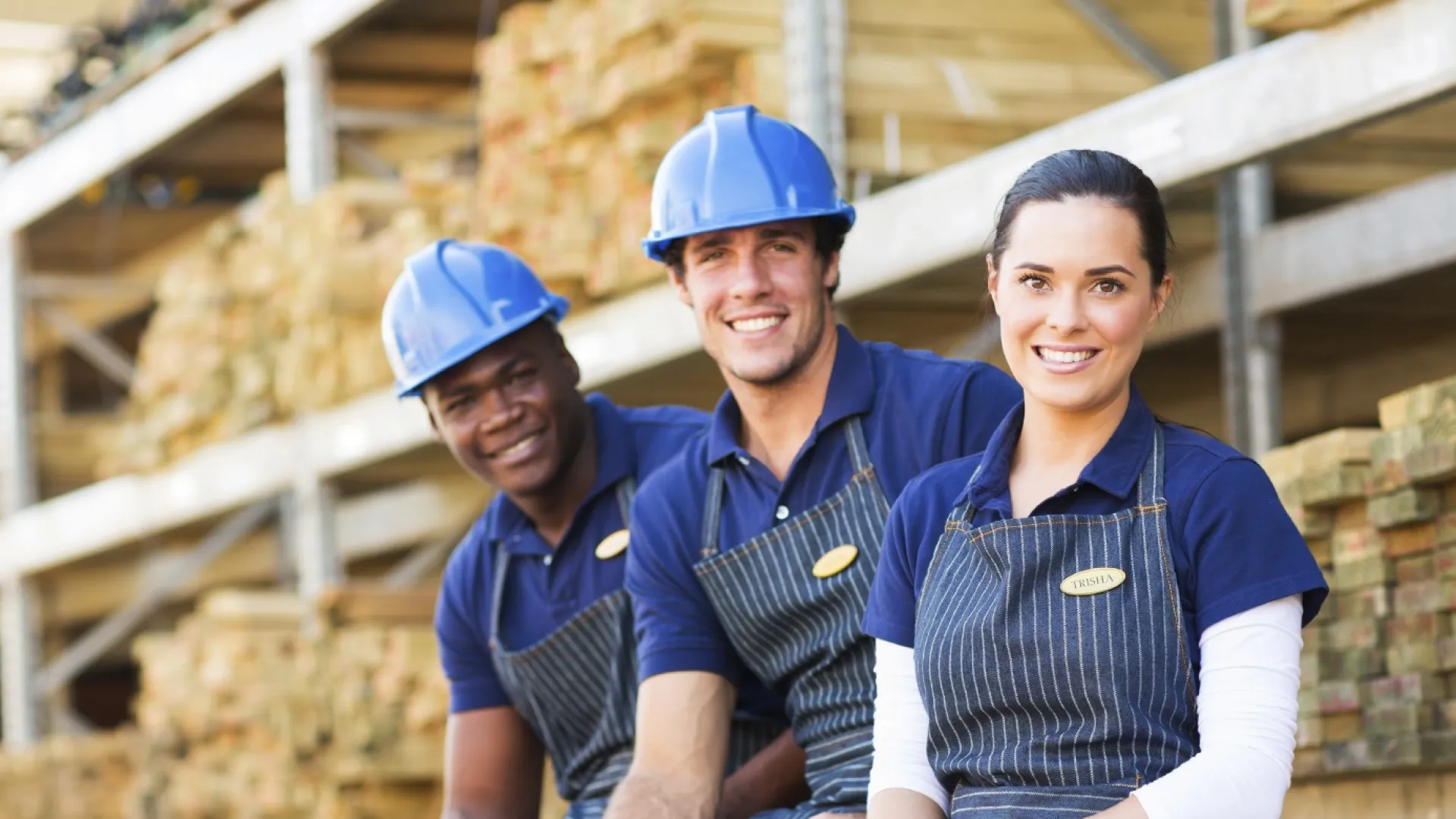 Three smiling workers in blue uniforms and safety helmets at a lumber yard with stacked wood in the background