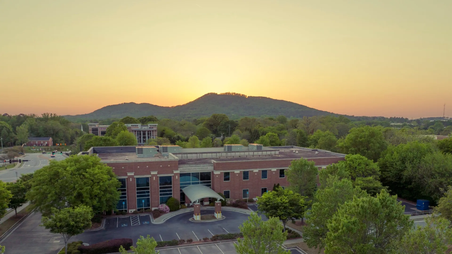 Aerial view of a brick office building surrounded by trees and empty parking lot at sunset with hills in the background.