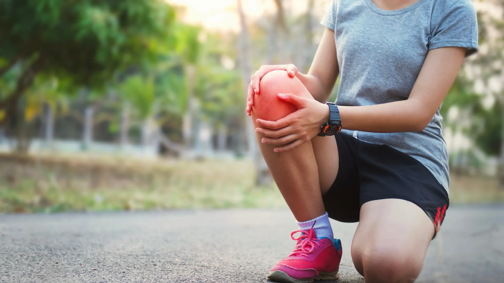 Person kneeling outdoors holding knee in pain, wearing sportswear and pink running shoes on pavement.