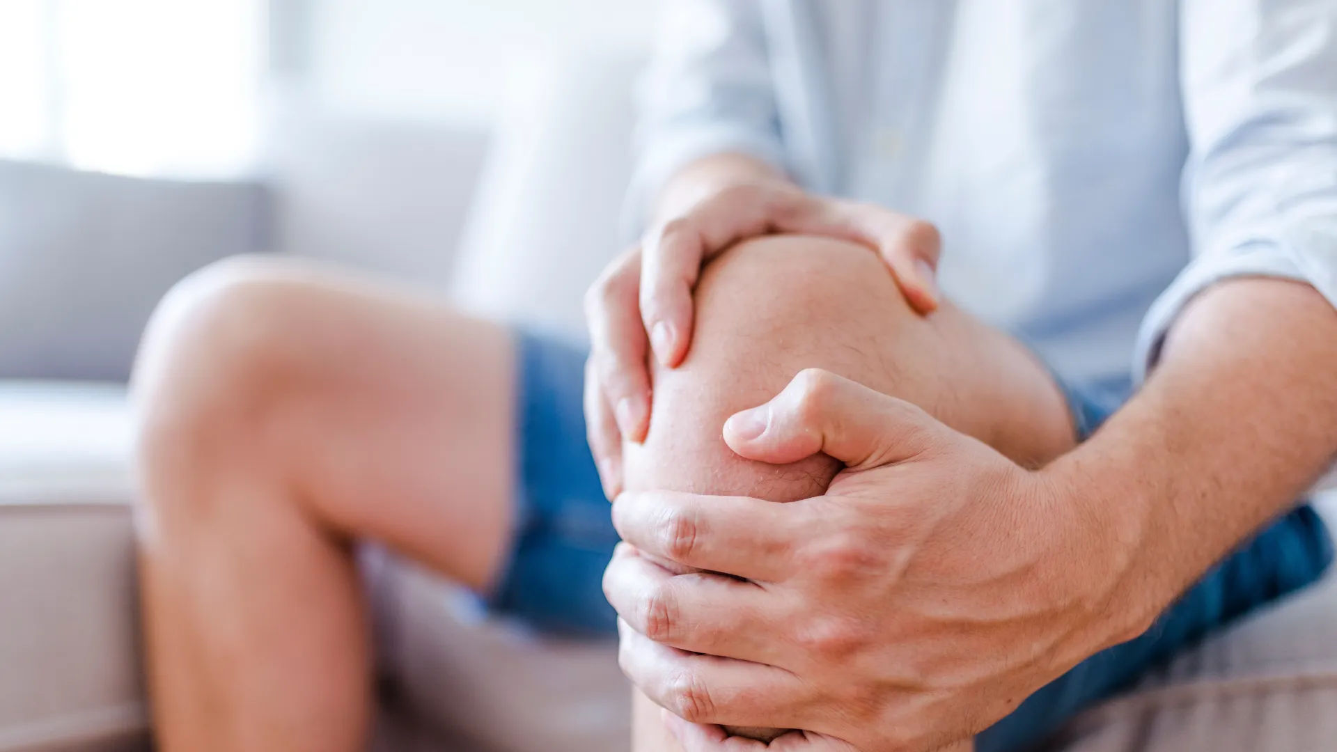 Person sitting on a couch holding their knee in pain indoors in casual shorts and shirt