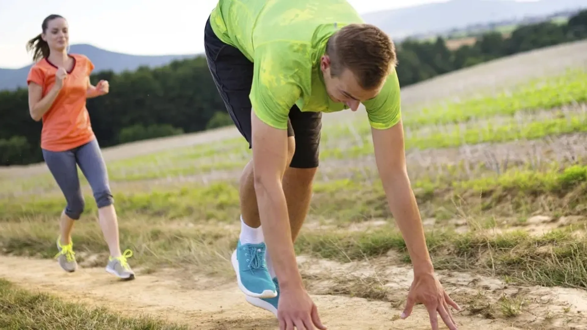 Runner in green shirt preparing to sprint on dirt path with a woman jogging behind in orange shirt under blue sky.