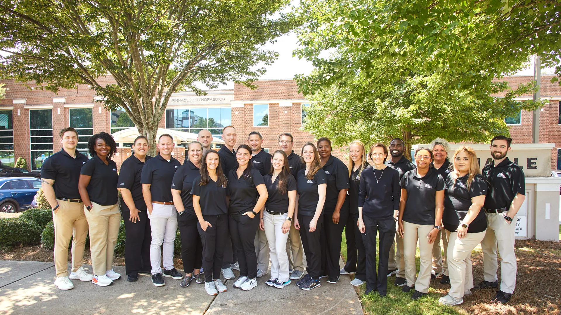 Group of diverse professionals in black shirts and beige or black pants posing outdoors near office building under trees.