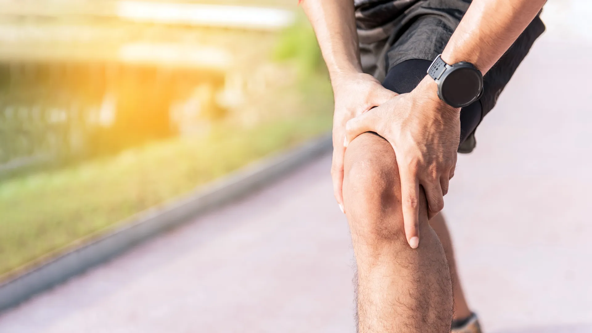 Runner holding knee in pain during outdoor exercise on sunny pathway, indicating joint or muscle discomfort.