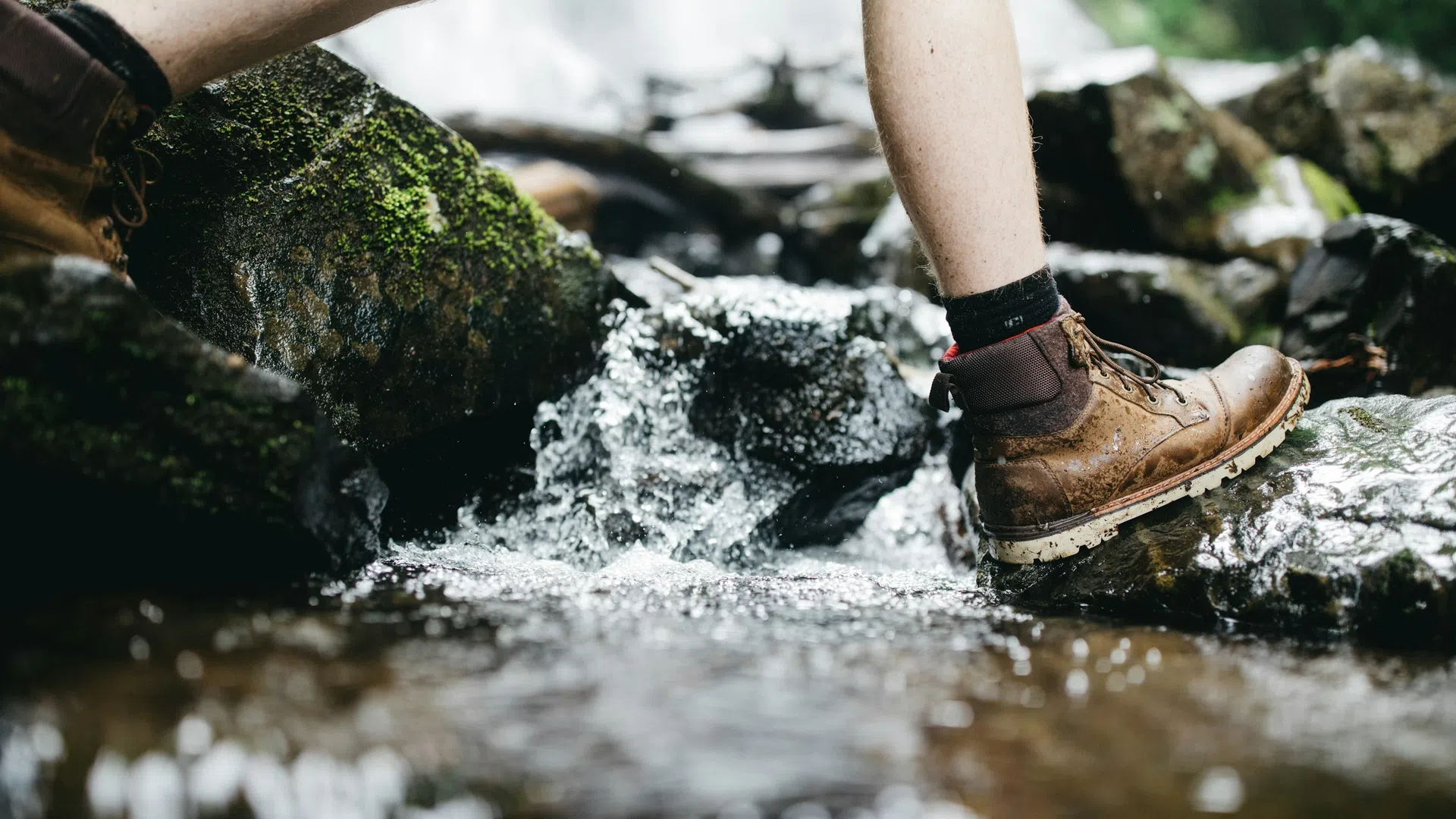 Close-up of a person wearing brown hiking boots crossing a rocky stream with flowing water and moss-covered stones.