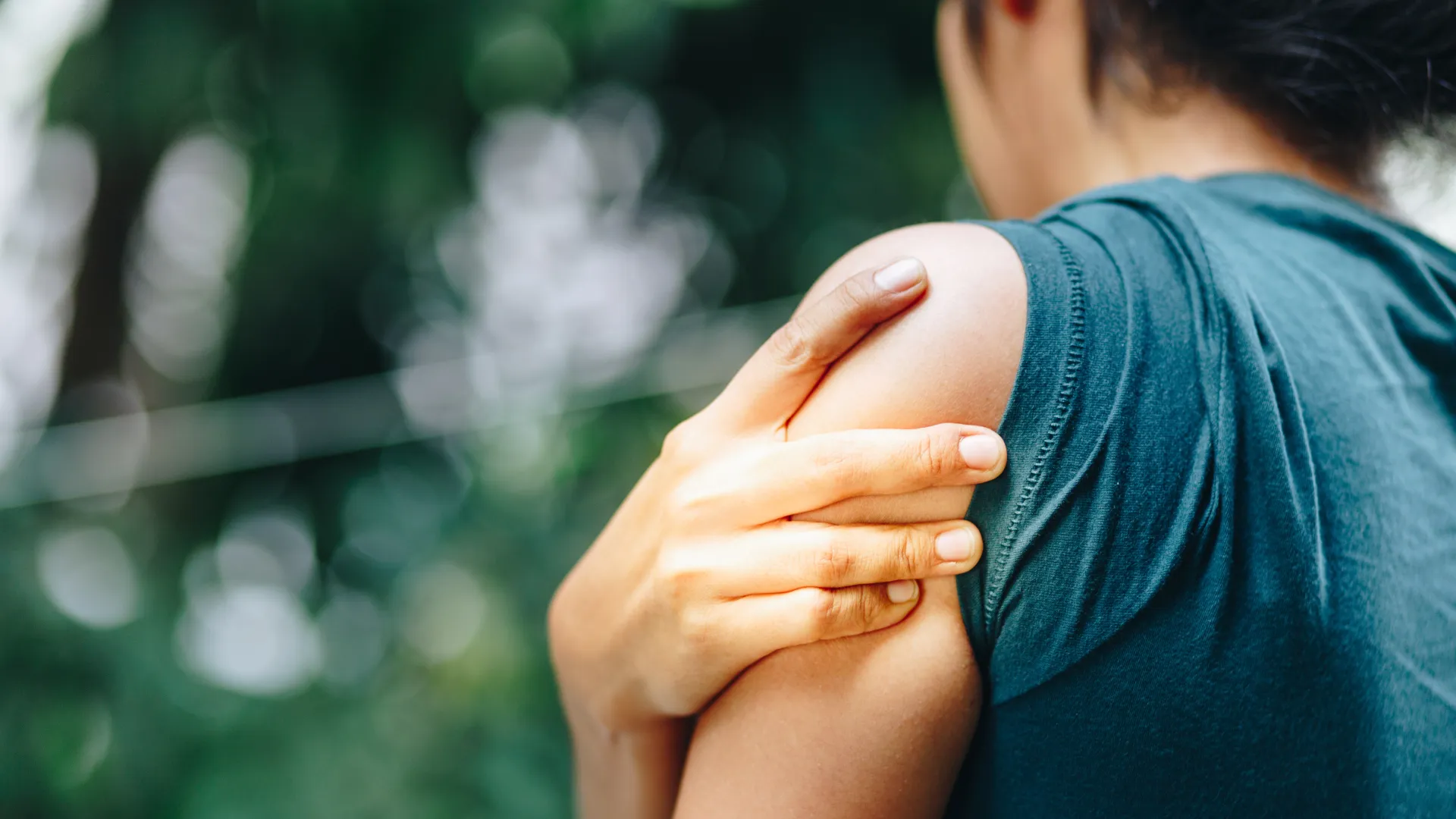 Person in teal shirt holding shoulder with hand, indicating shoulder pain or discomfort outdoors.