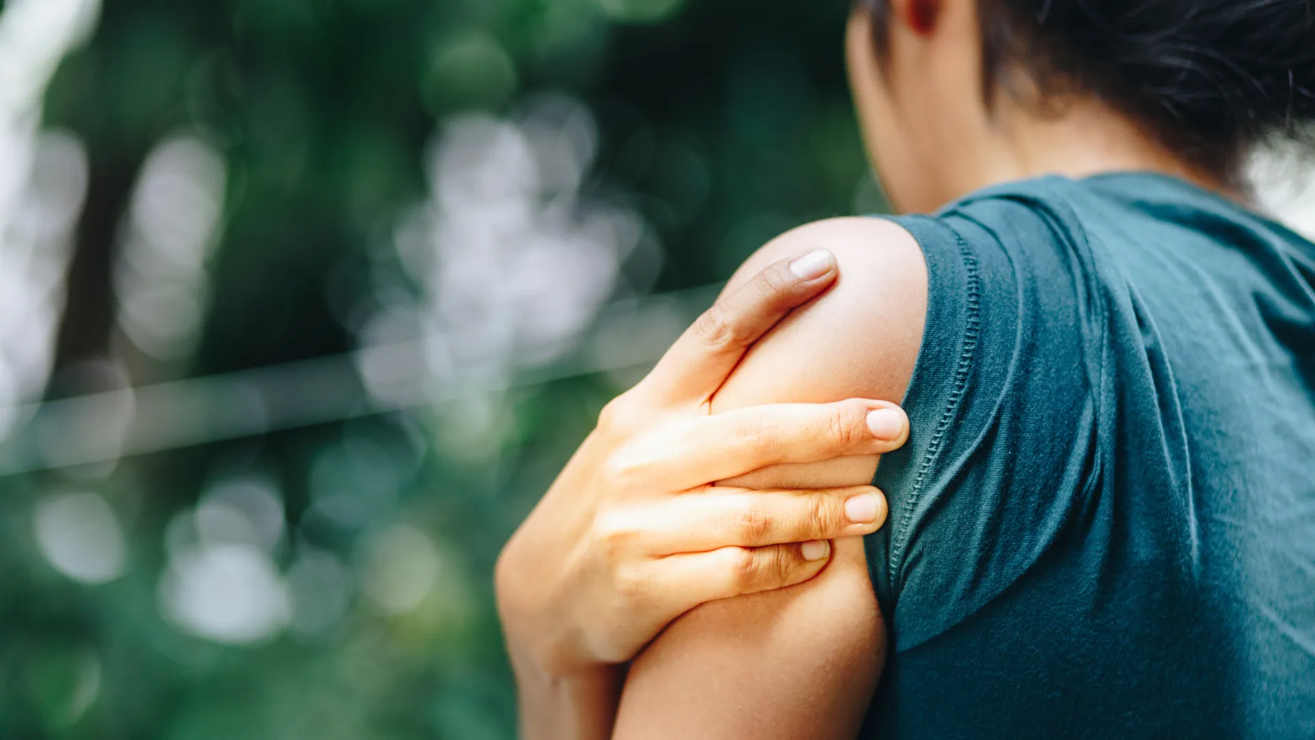 Person in teal shirt holding shoulder with hand, indicating shoulder pain or discomfort outdoors.