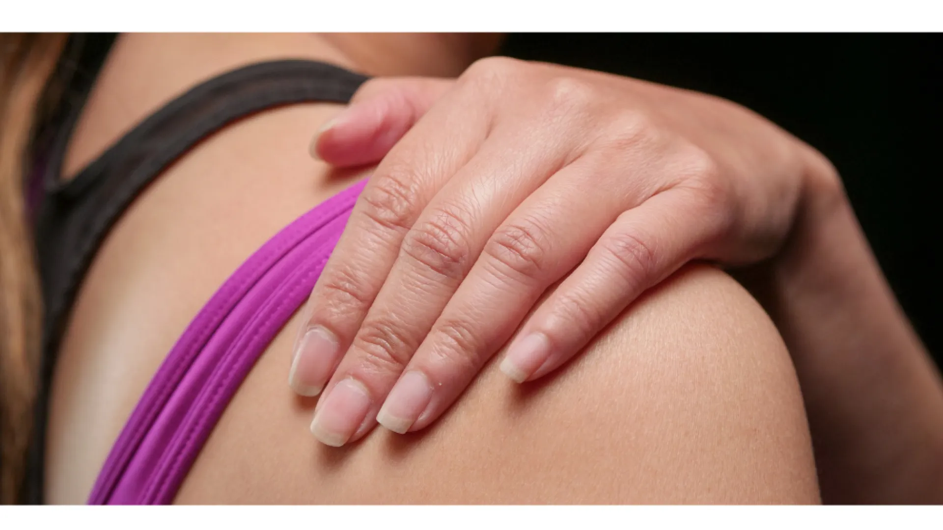 Close-up of a person touching their shoulder, wearing a purple and black sleeveless top, against a dark background
