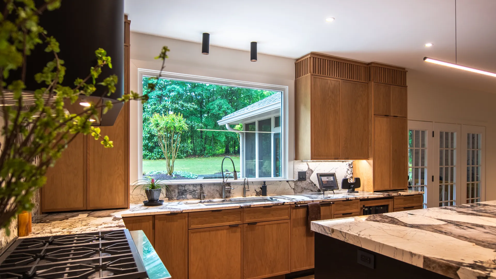 Modern kitchen with wooden cabinets, marble countertops, large window view of green backyard, and stainless steel stove.