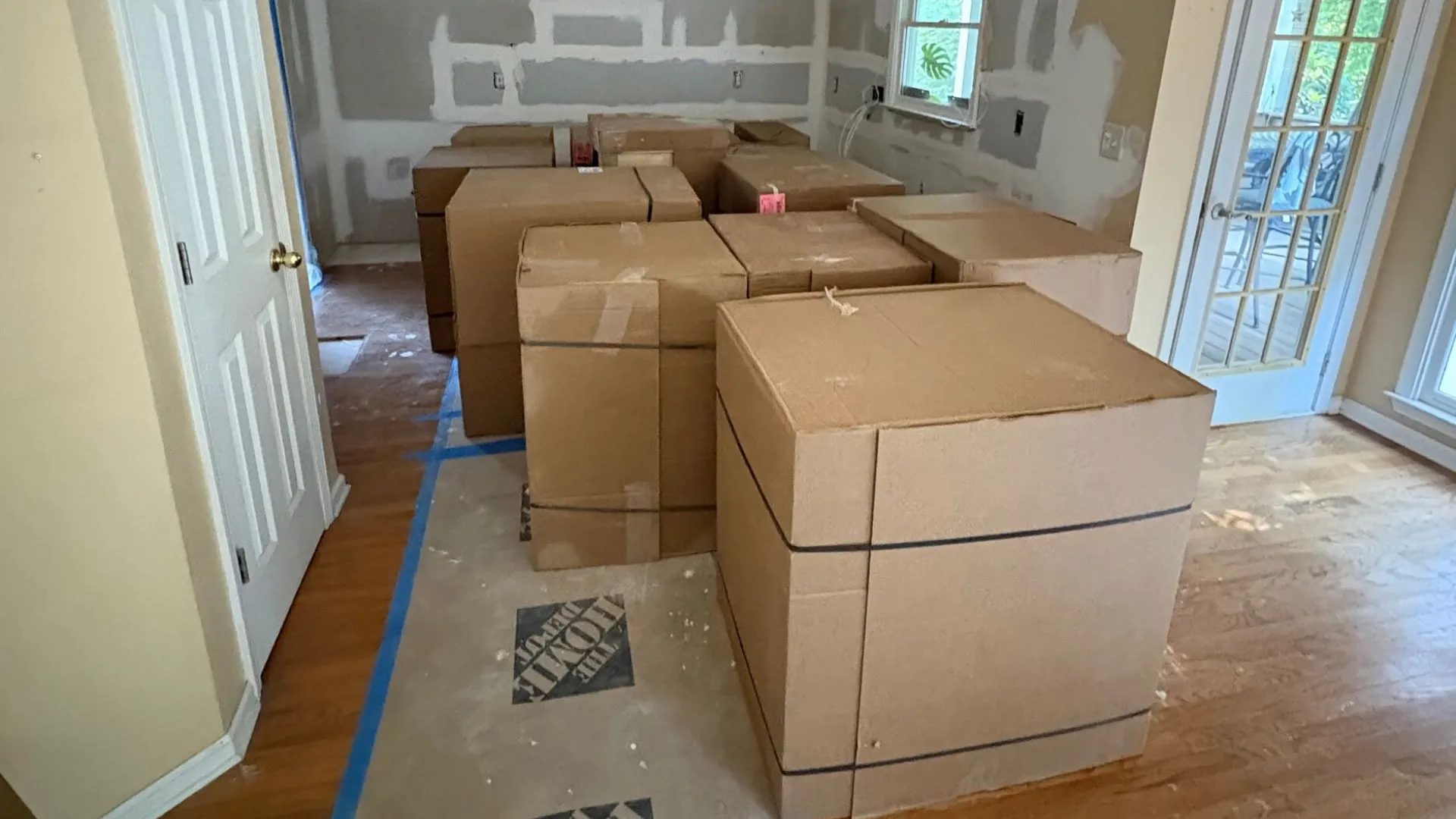 Packed cardboard boxes in a partially renovated room with drywall patches and hardwood floors.