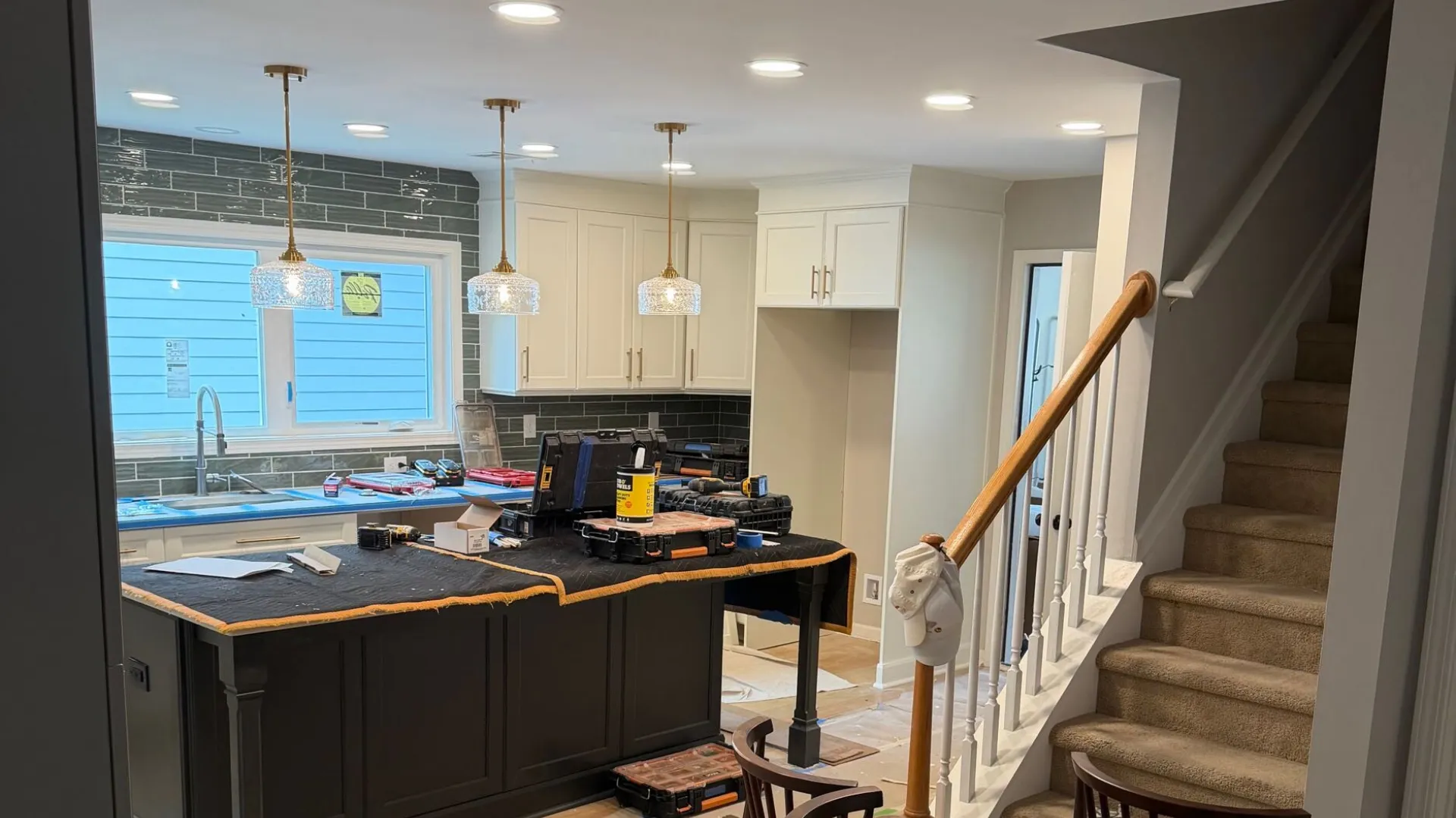Modern kitchen under renovation with white cabinets, black island, wooden chairs, and carpeted staircase.