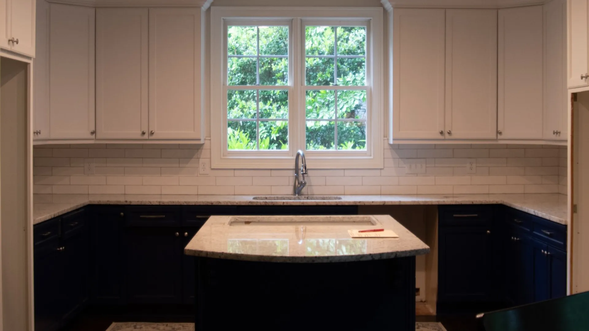 Modern kitchen with white upper cabinets, navy blue lower cabinets, center island, and large window with greenery view