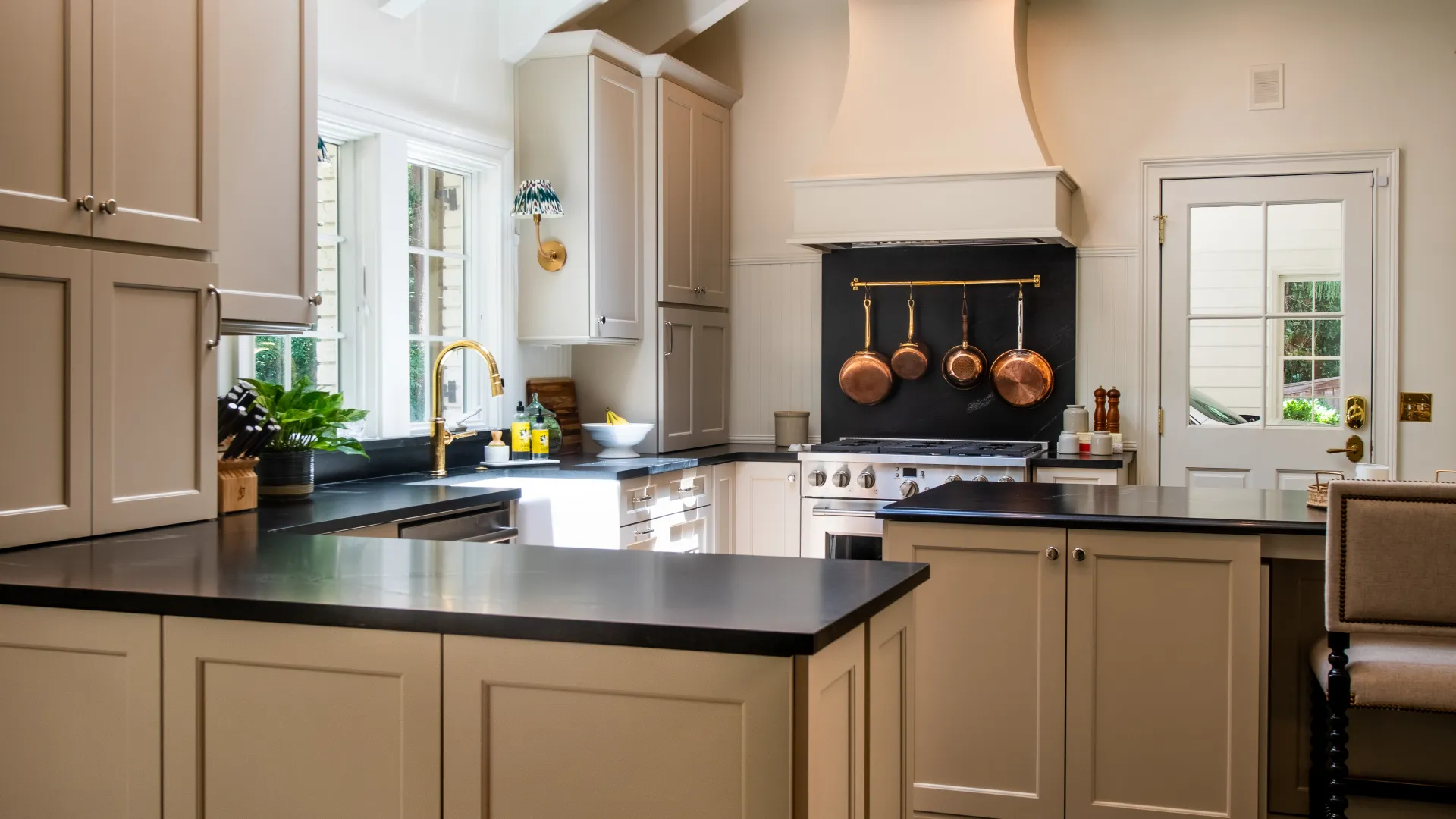 Bright modern kitchen with beige cabinets, black countertops, hanging copper pots, and a large window.