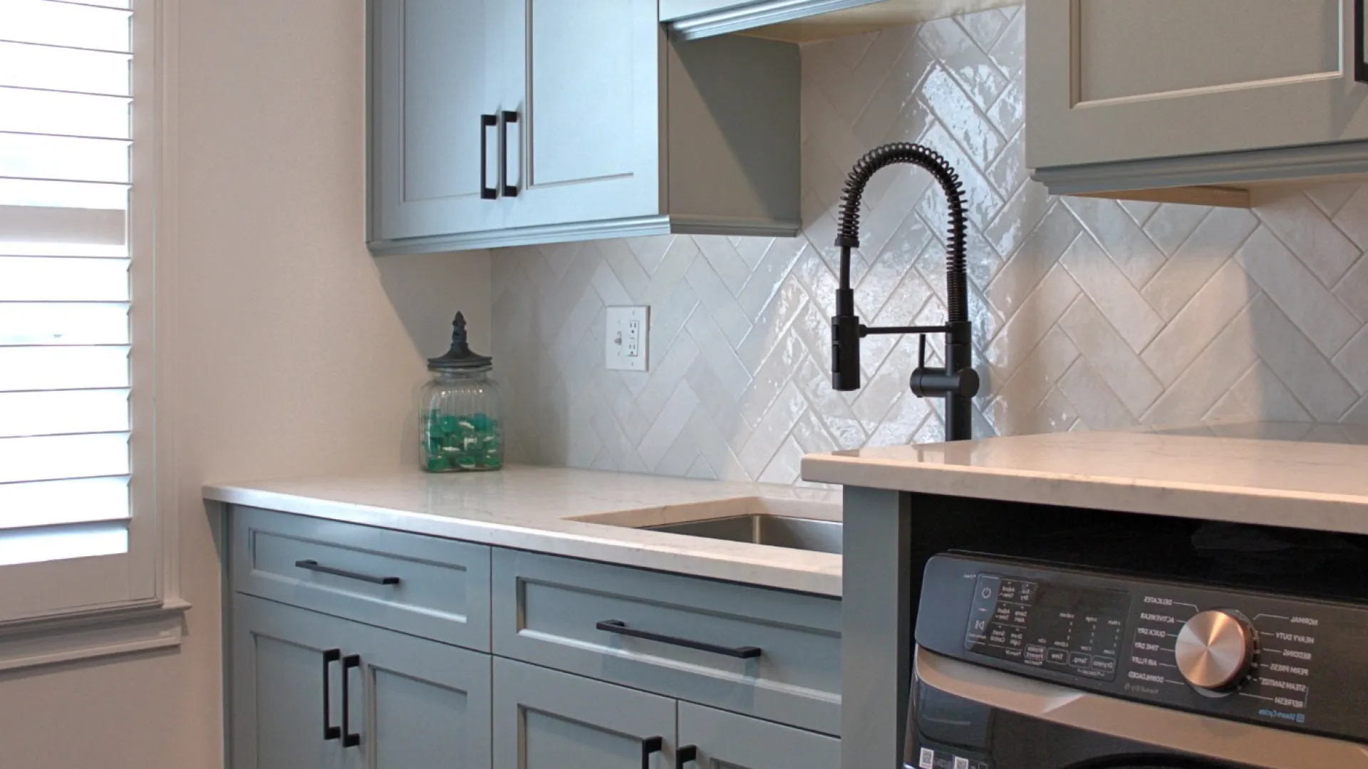 Modern laundry room with gray cabinets, black faucet, white countertops, backsplash, and stainless steel washer dryer.