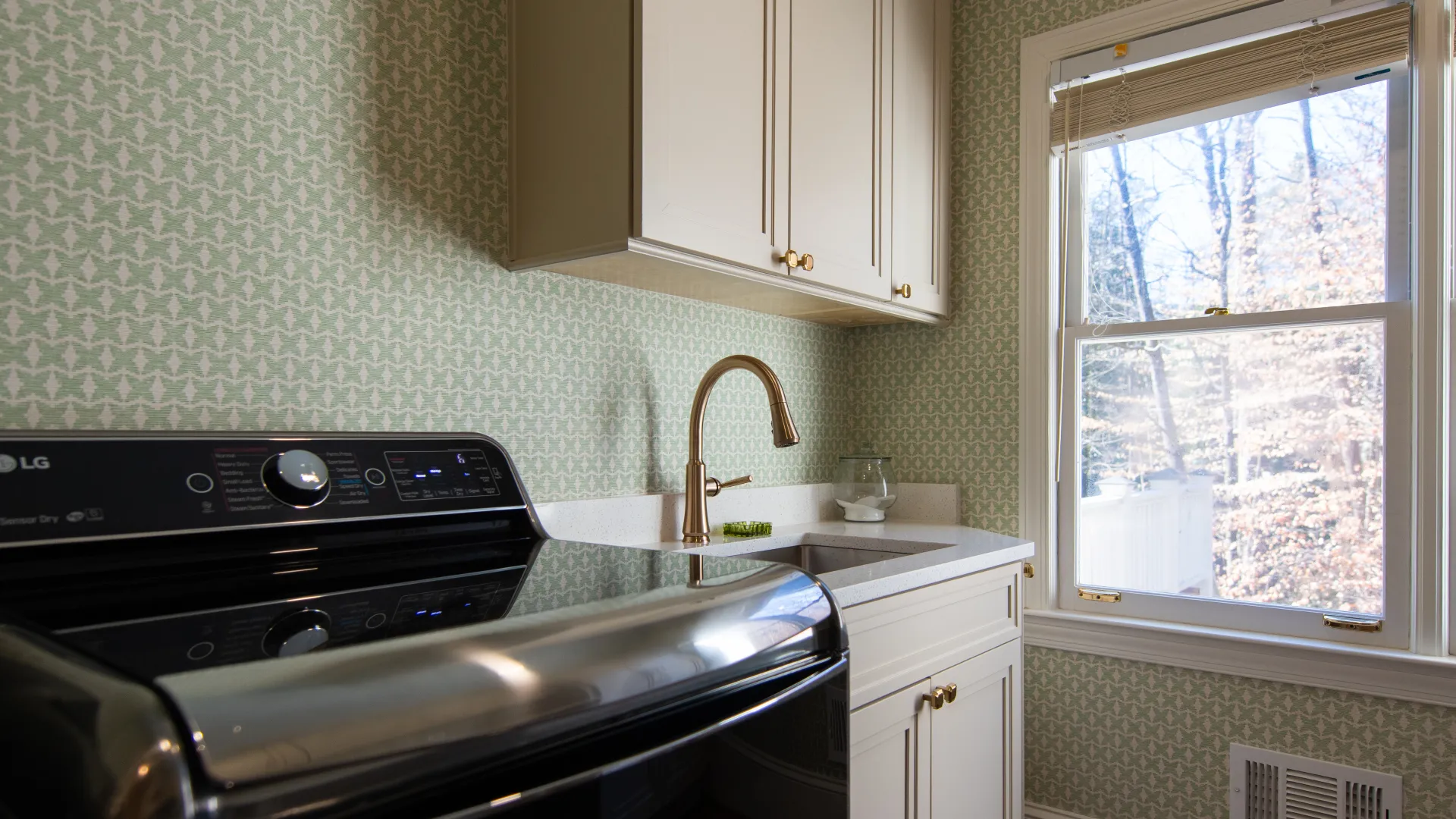 Modern laundry room with black LG washer dryer, gold faucet, white cabinets, and patterned green wallpaper.