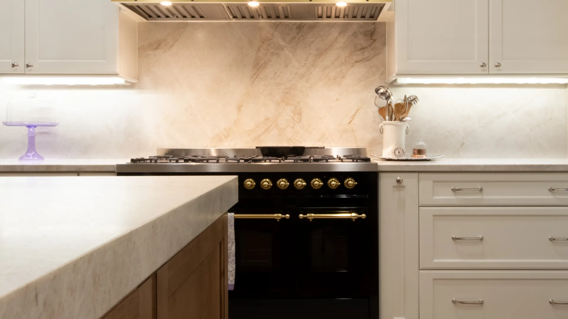 Modern kitchen with white cabinets, beige marble backsplash, black stove with gold accents, and wooden island.