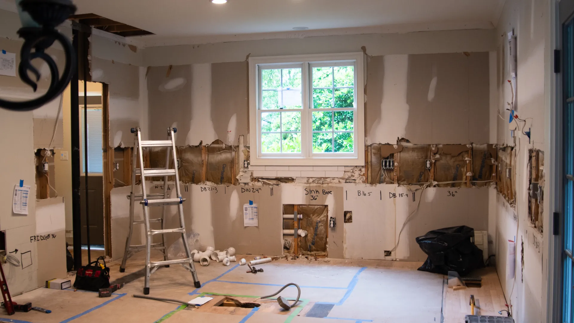 Kitchen under renovation with exposed walls, insulation, pipes, ladder, and tools under a window.