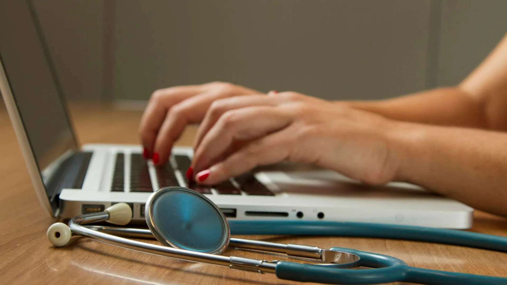 Hands typing on a laptop keyboard with a stethoscope resting on the wooden table nearby, symbolizing healthcare and technology.