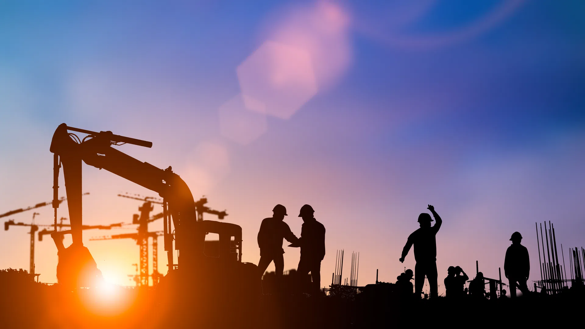 Silhouettes of construction workers and machinery at sunset with cranes in background and blue sky.