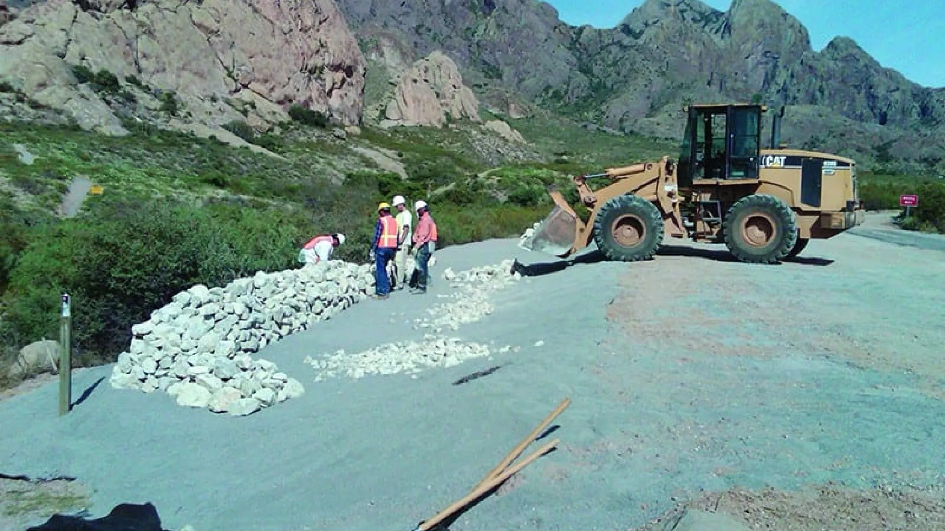 Construction workers building a stone wall with heavy machinery in a mountainous outdoor setting on a clear day.