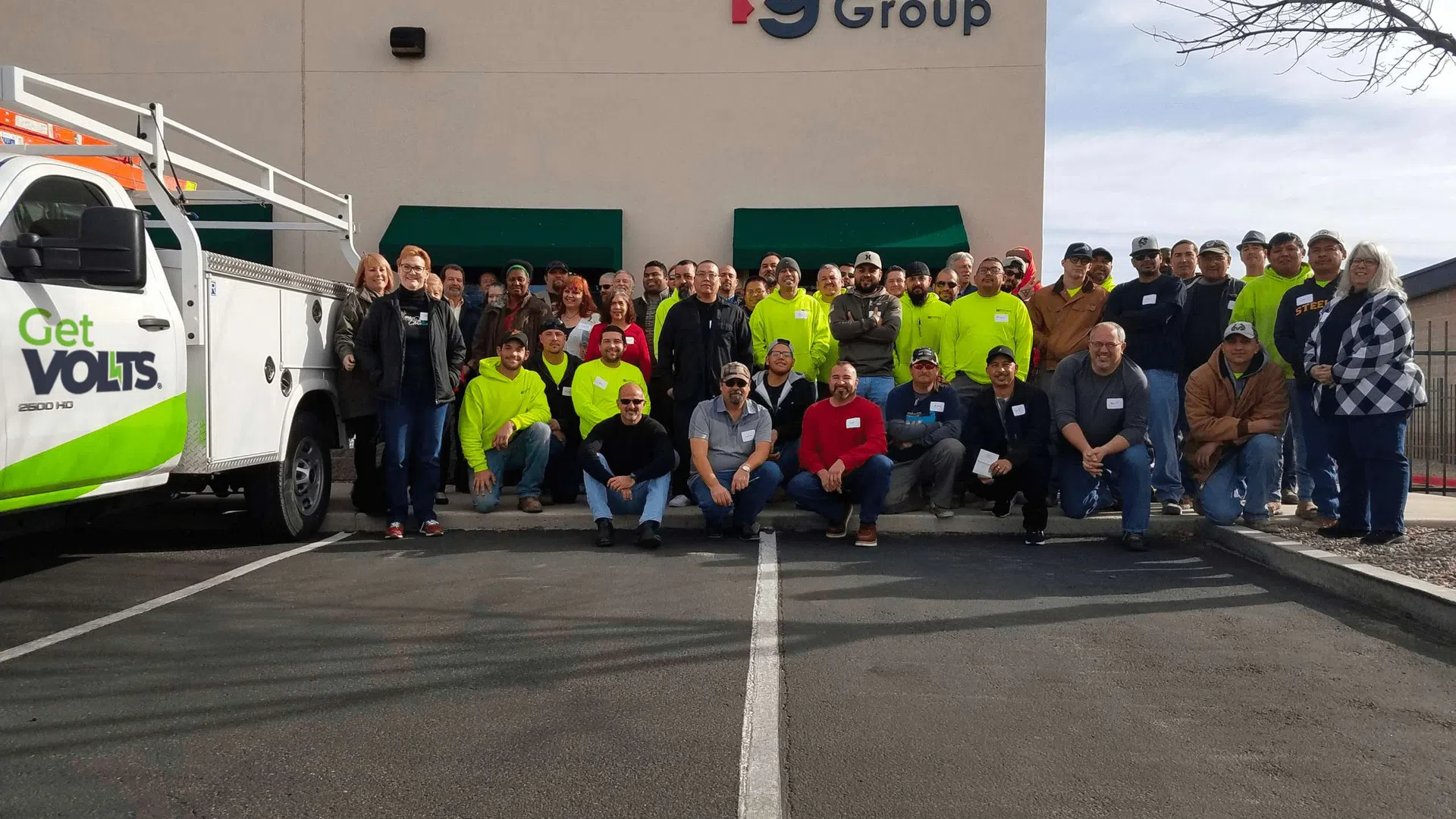Group of workers posing outside Burgos Group building with a Get Volts service truck parked nearby under clear sky.