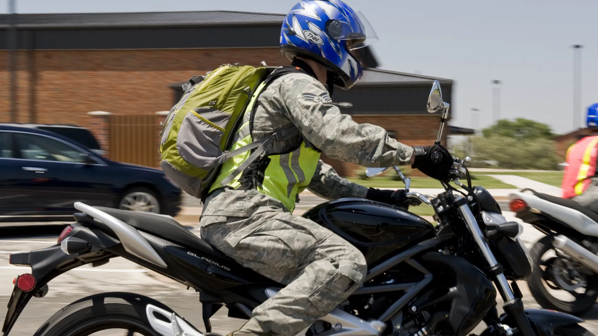Motorcyclist in military uniform with blue helmet and green backpack riding a black motorcycle in an urban area.