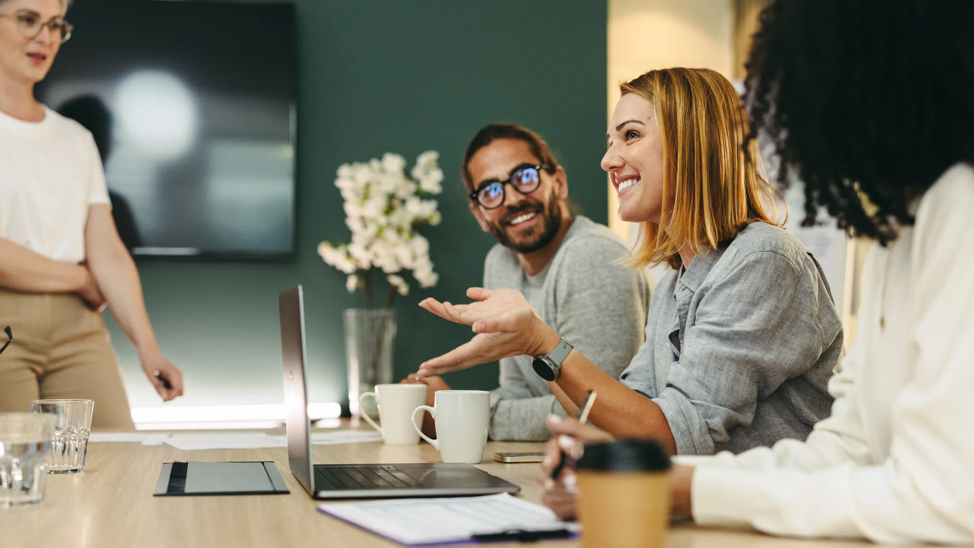 Diverse coworkers engaged in a lively meeting around a conference table with laptops and coffee cups.