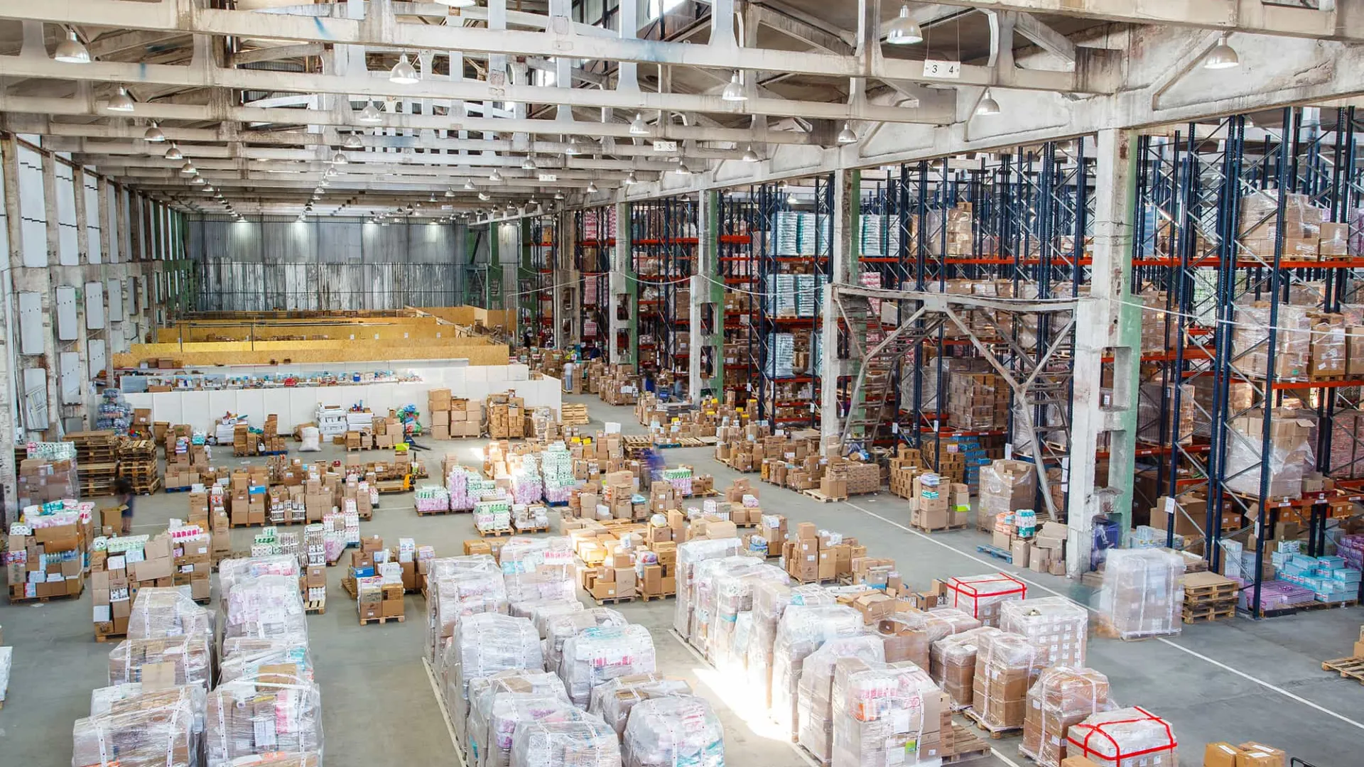 Large warehouse filled with stacked pallets and boxes under white metal truss ceiling with bright lighting