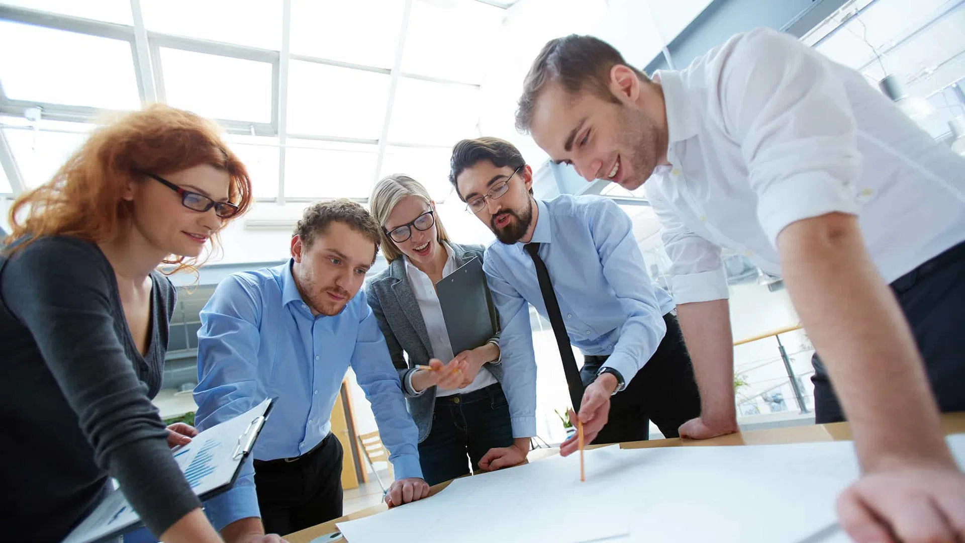 Group of five professionals collaborating over large documents on a table in a bright office.