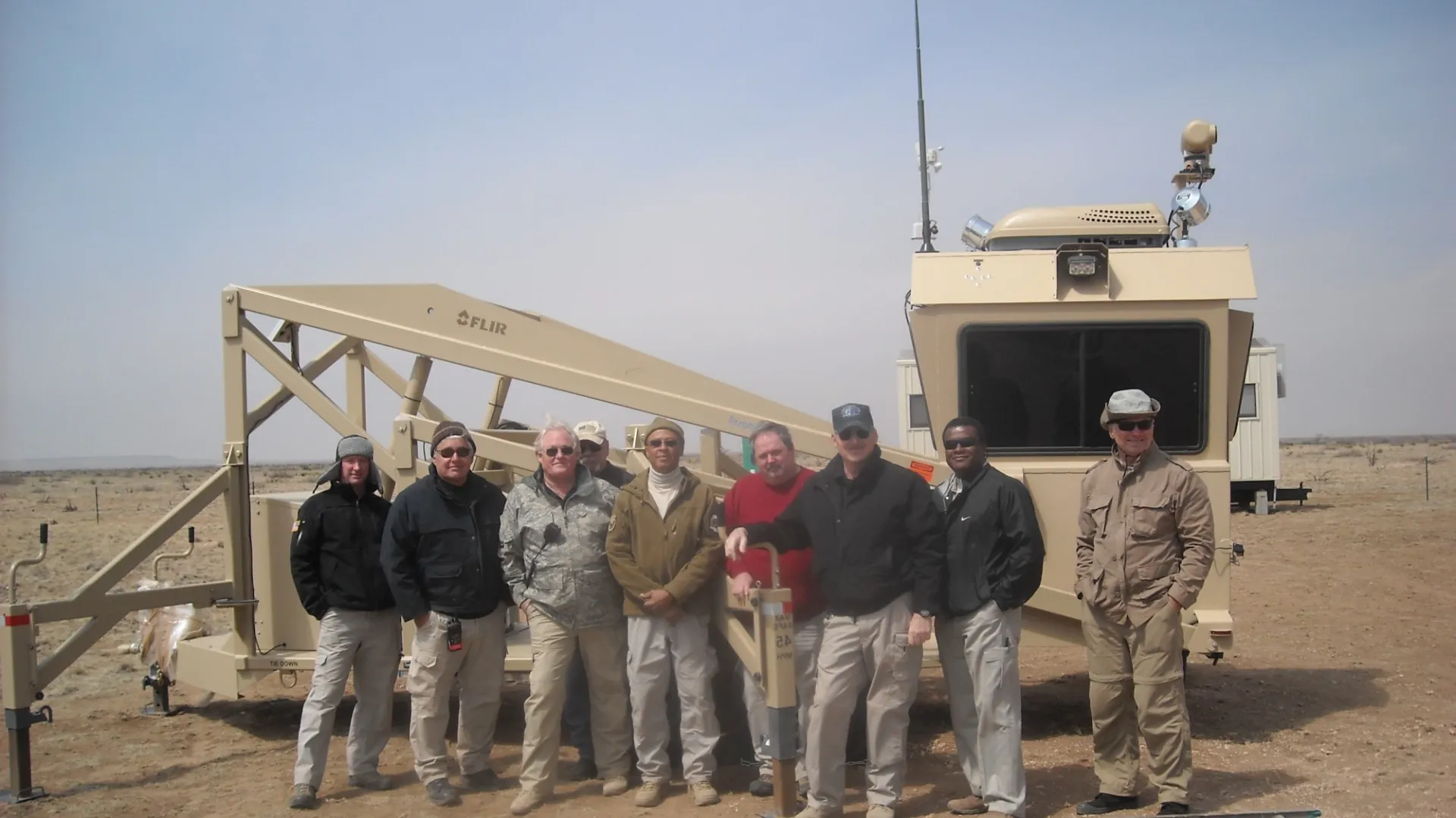 Group of eight people standing in front of large beige industrial equipment on a barren outdoor site.