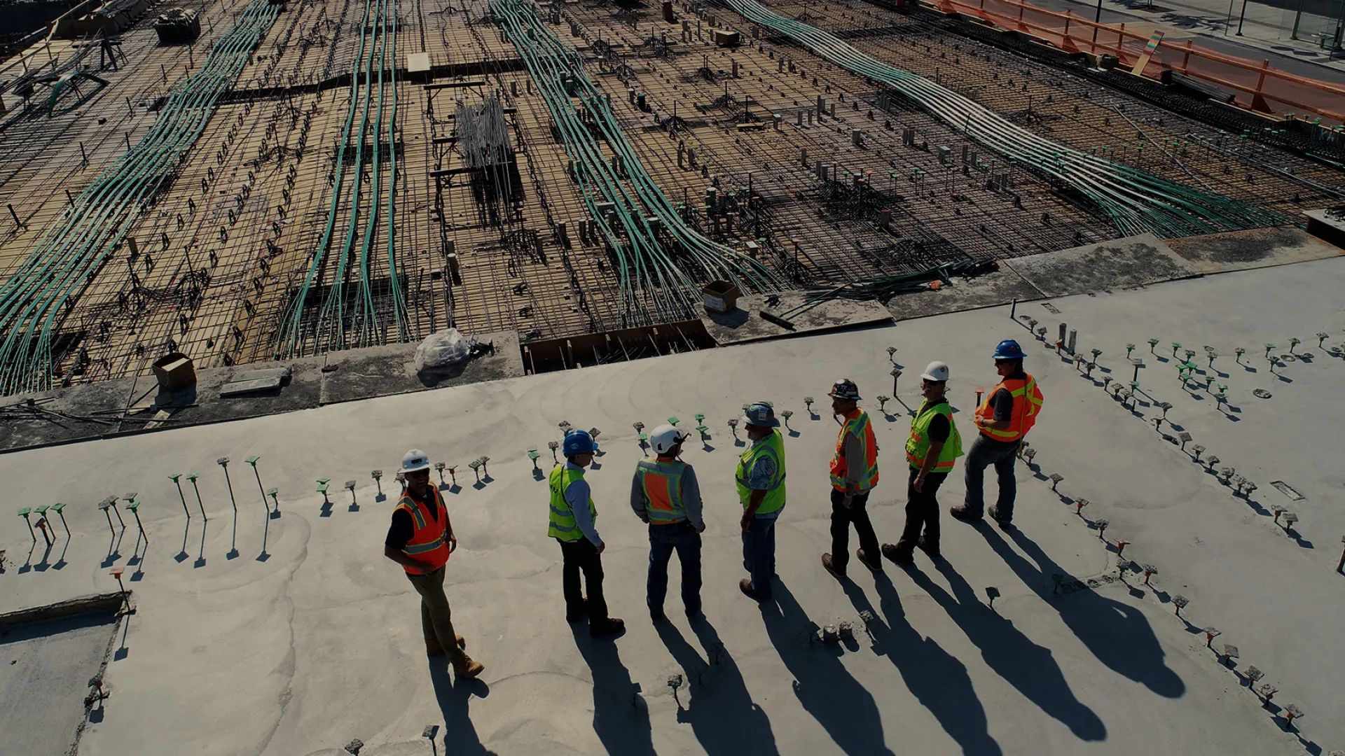 Construction workers in safety gear inspect a large concrete slab with visible plumbing pipes and rebar below.