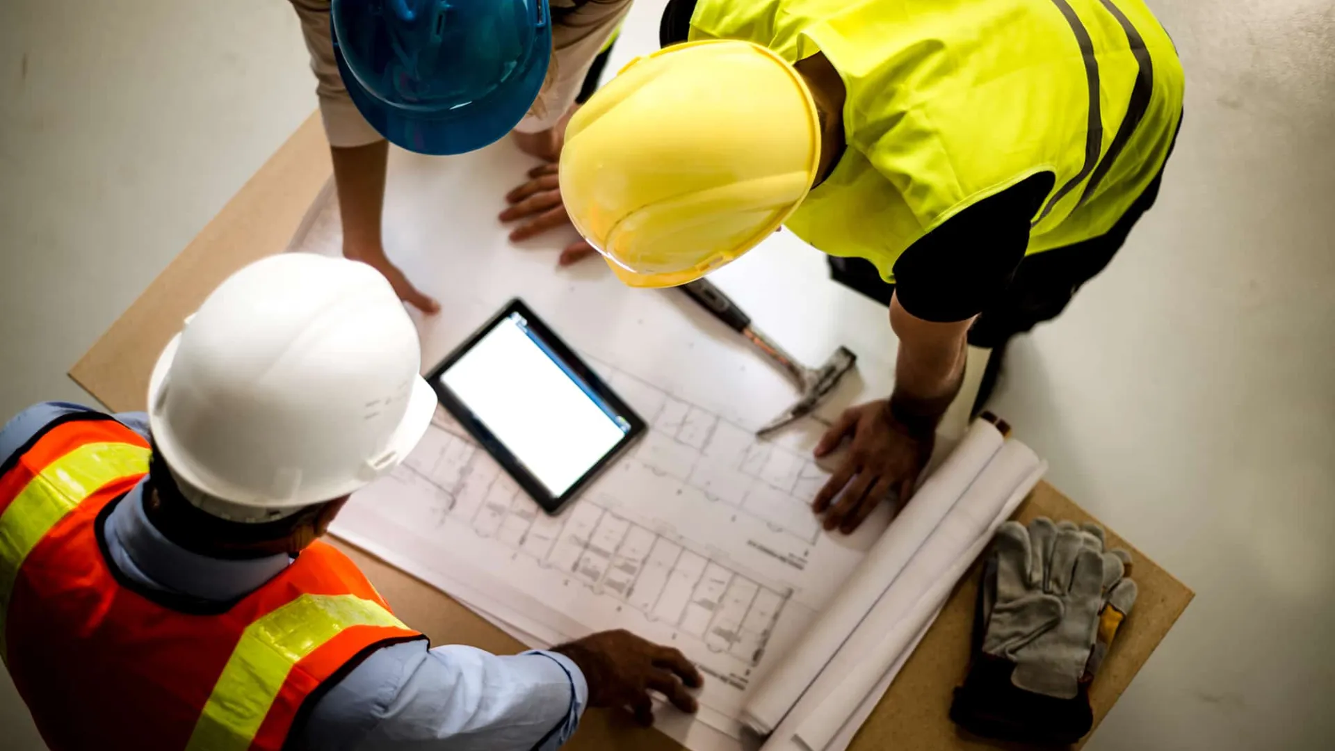 Top view of construction workers wearing helmets reviewing blueprints and digital tablet at worksite.