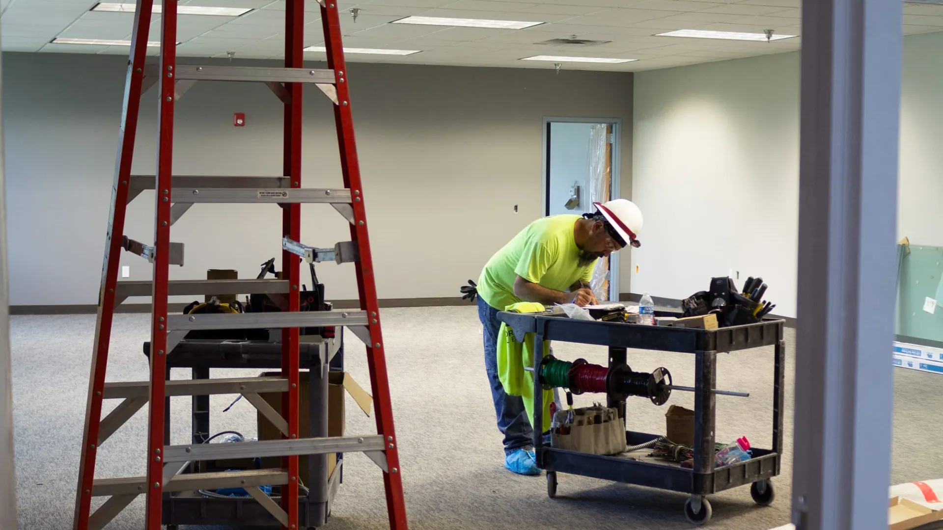 Construction worker in neon shirt and hard hat checking notes beside equipment cart and red ladder in office space.