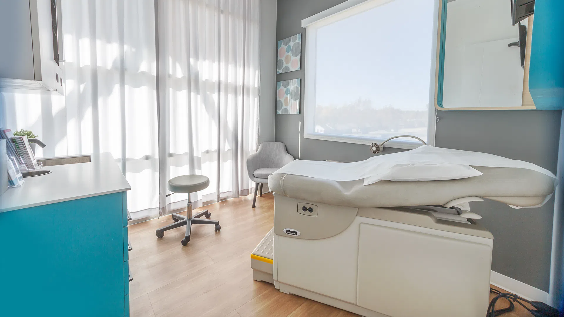 Bright medical examination room with adjustable patient bed, stool, chair, and blue cabinets under natural light.