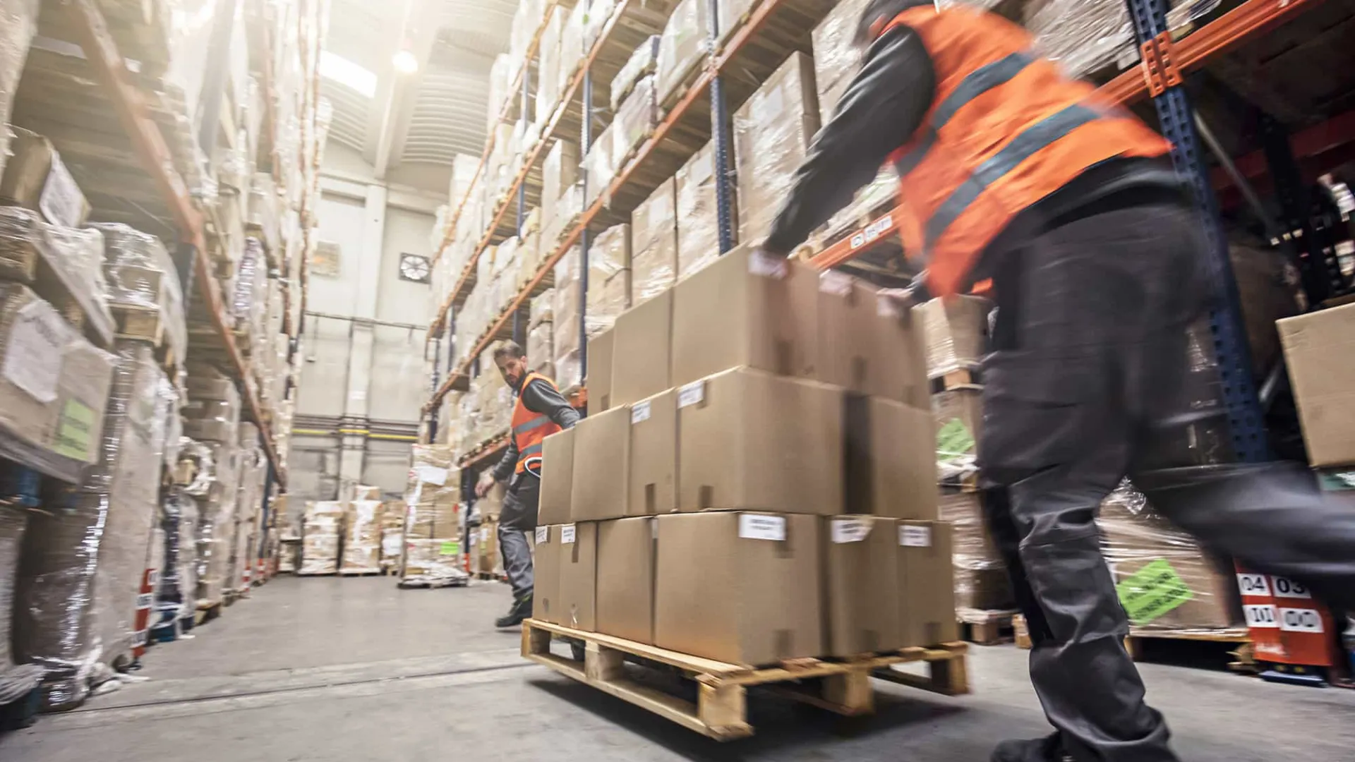 Warehouse workers wearing orange vests moving boxes on pallets in a large storage facility with tall shelves.