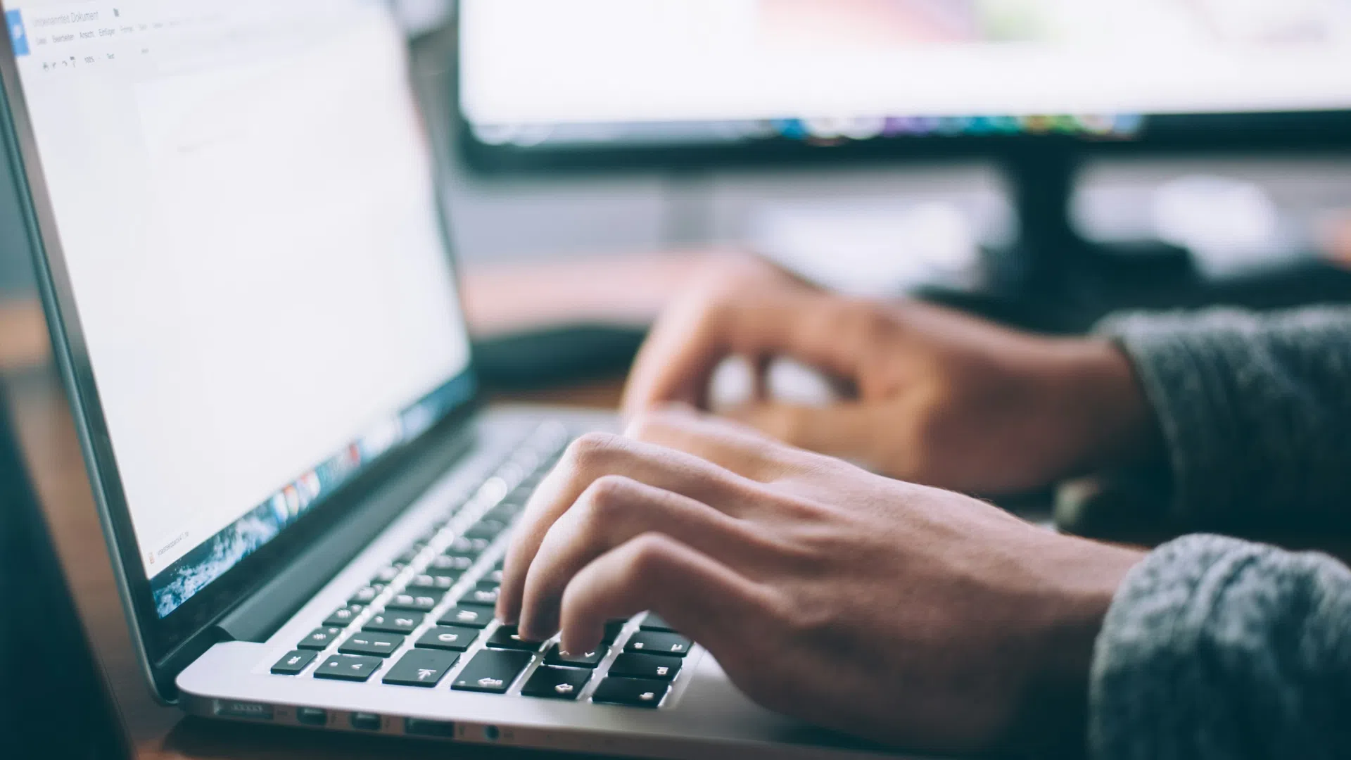 Close-up of hands typing on a laptop keyboard with a blurred monitor and workspace in background