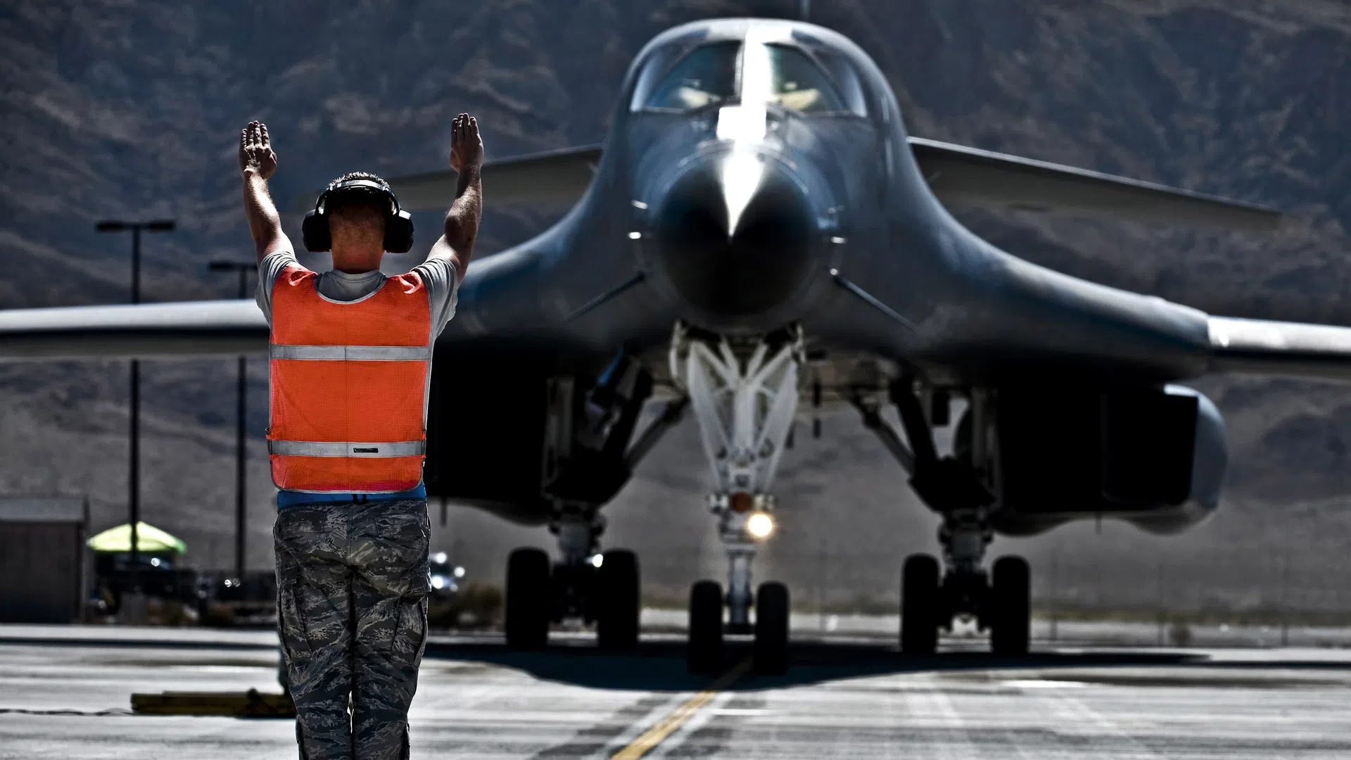 Ground crew member directing a large military aircraft on a runway in a mountainous desert area.