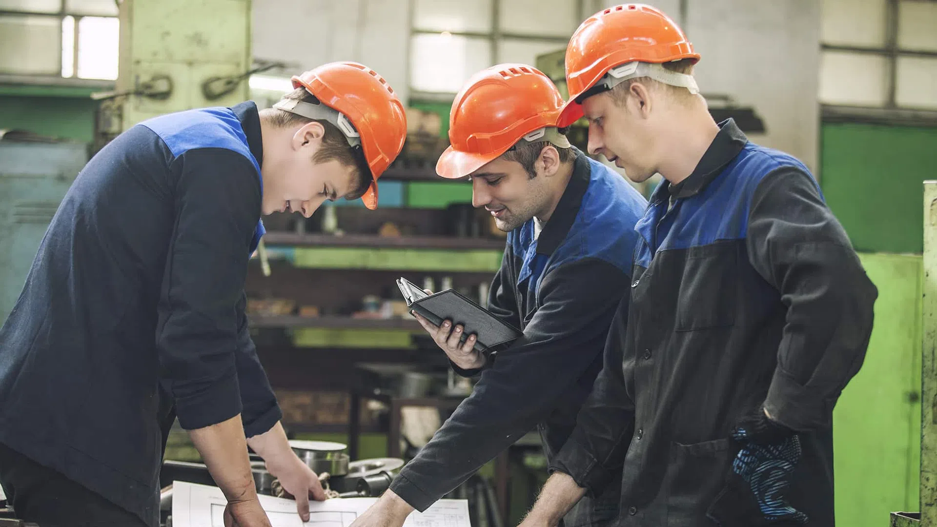 Three male factory workers in orange helmets reviewing blueprints and tablet in industrial workshop