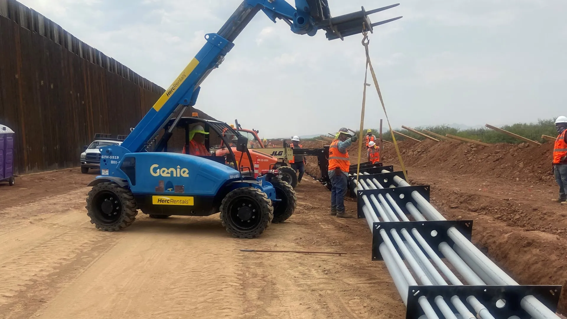 Blue Genie forklift moving large metal pipes at a construction site with workers in safety gear.