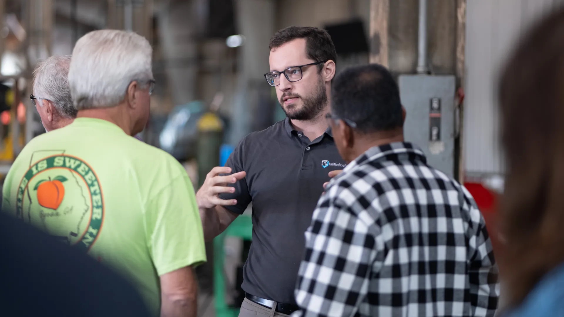 Man in glasses explaining something to a small group inside an industrial workshop with machinery in the background.