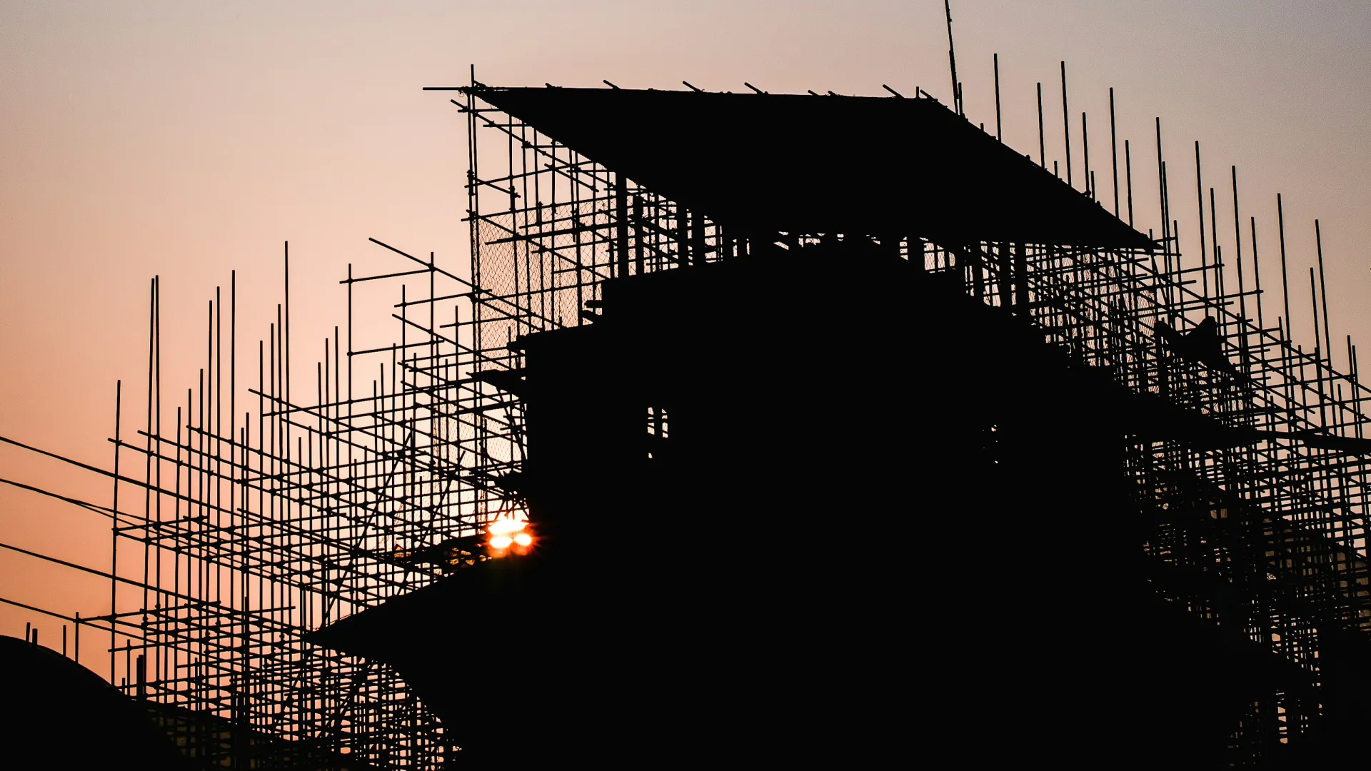 Silhouette of scaffolding and building structure against a gradient sunset sky with sun flare.