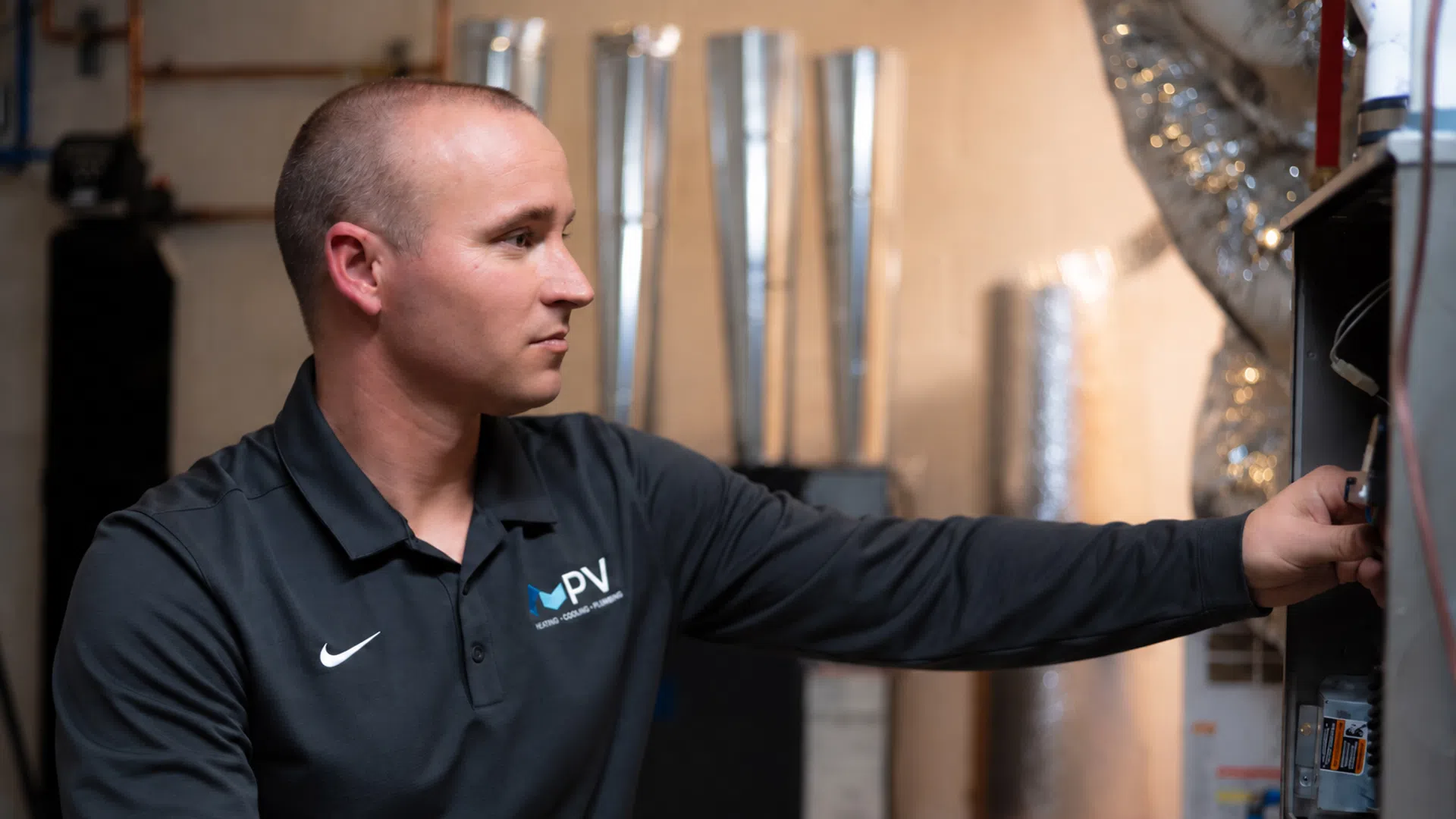 Technician in black PV Energy Solutions shirt inspecting HVAC or electrical system in a utility room.