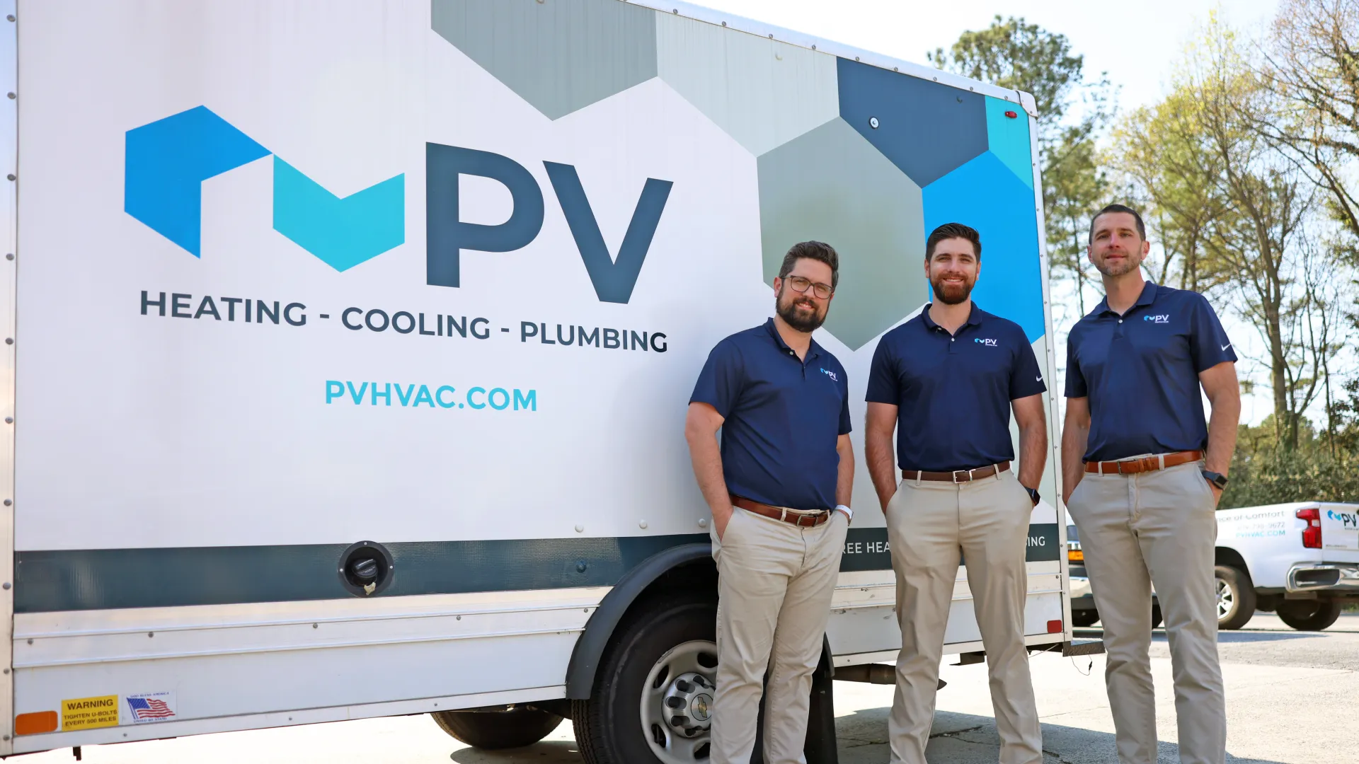 Three men in matching uniforms stand in front of a PV Heating Cooling Plumbing service truck outdoors.