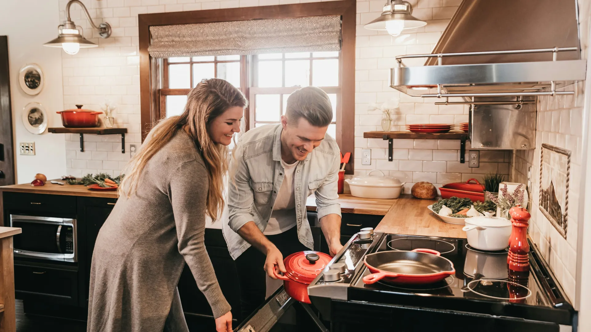 Couple cooking together in a modern kitchen, placing a red pot into the oven with smiles and warm lighting.
