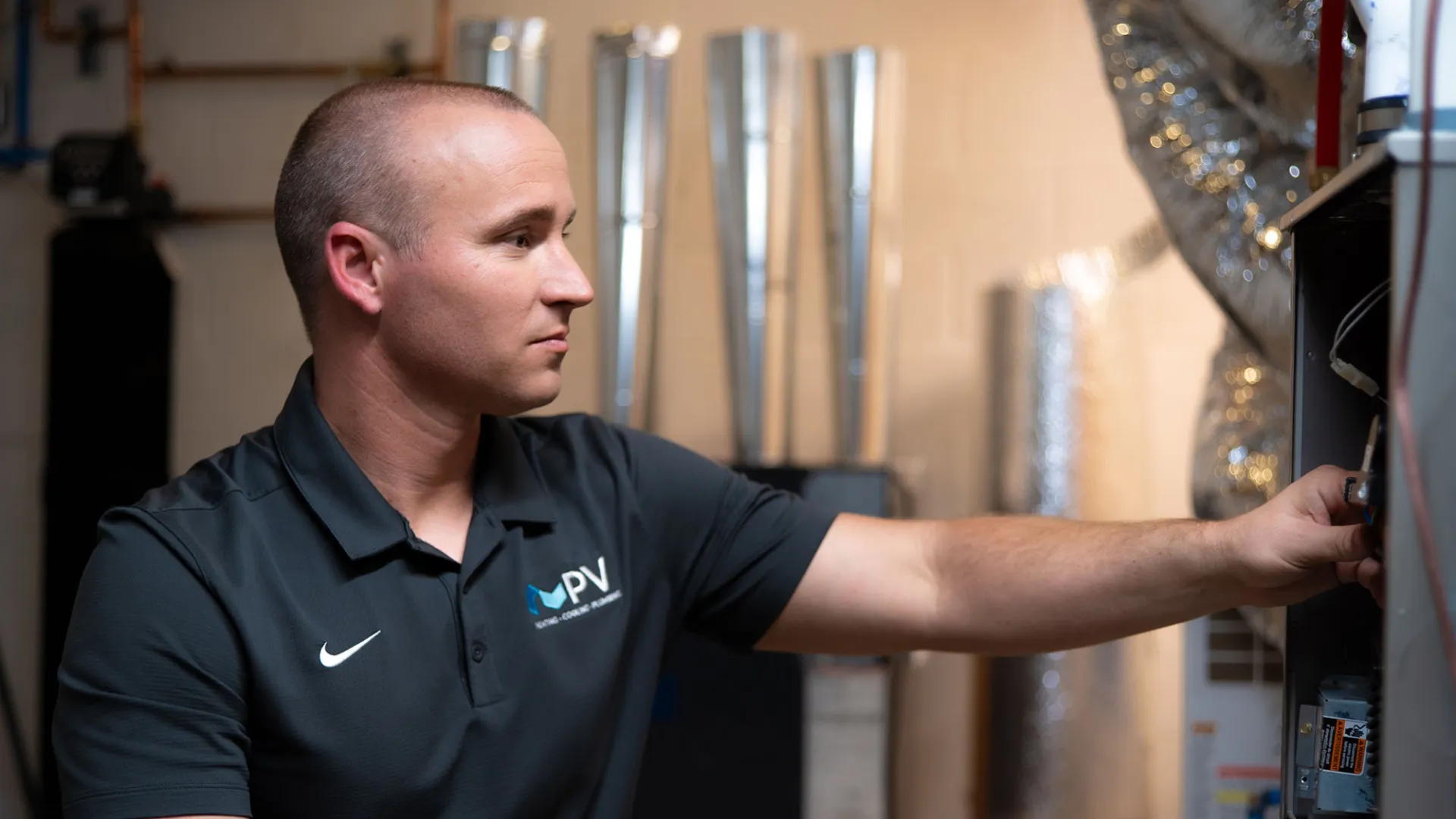 Technician in PV uniform adjusting HVAC system components in a utility room with metal ducts and pipes.