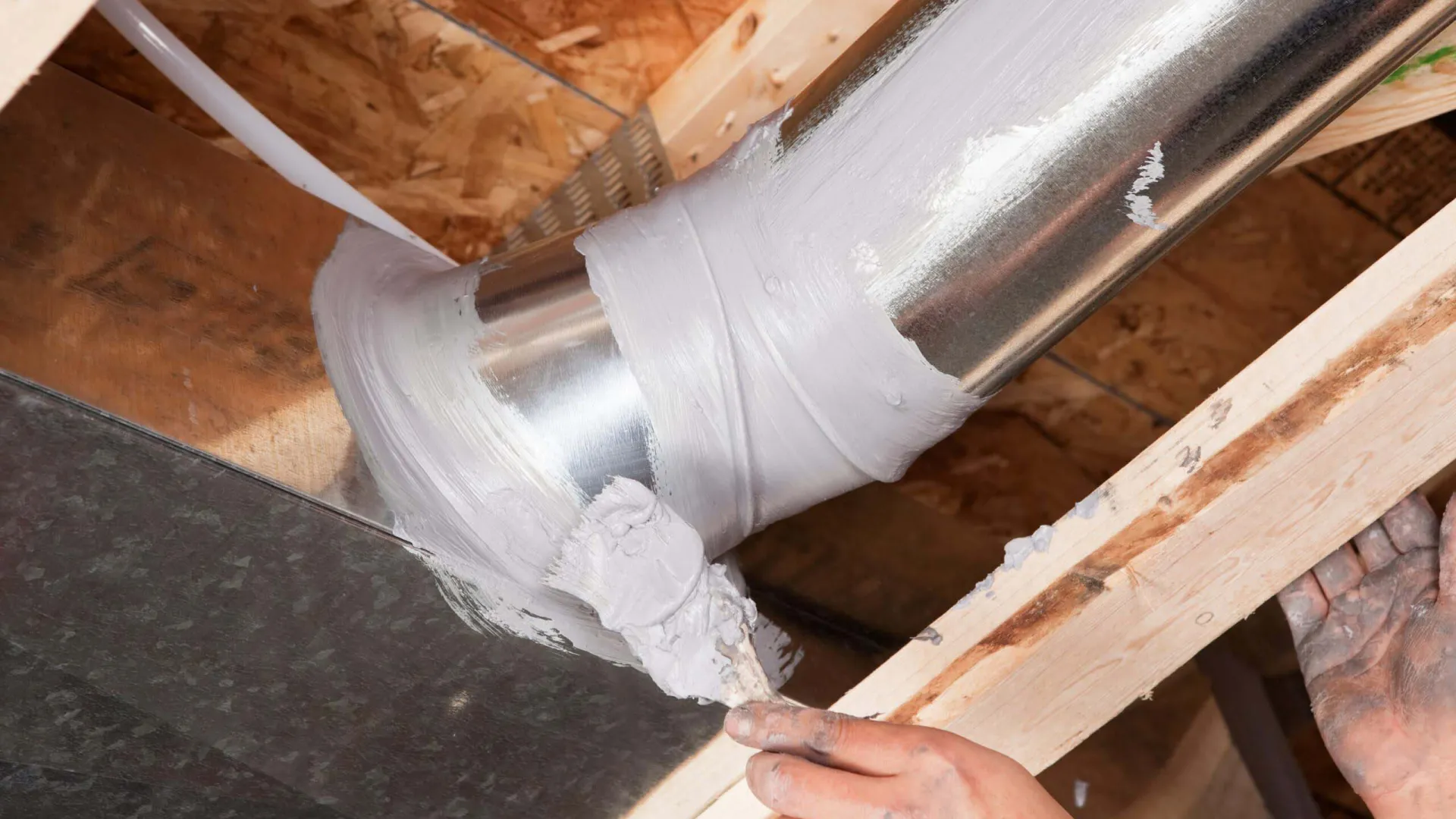 Hand applying white fireproof sealant around a metal duct pipe in a wooden ceiling frame.