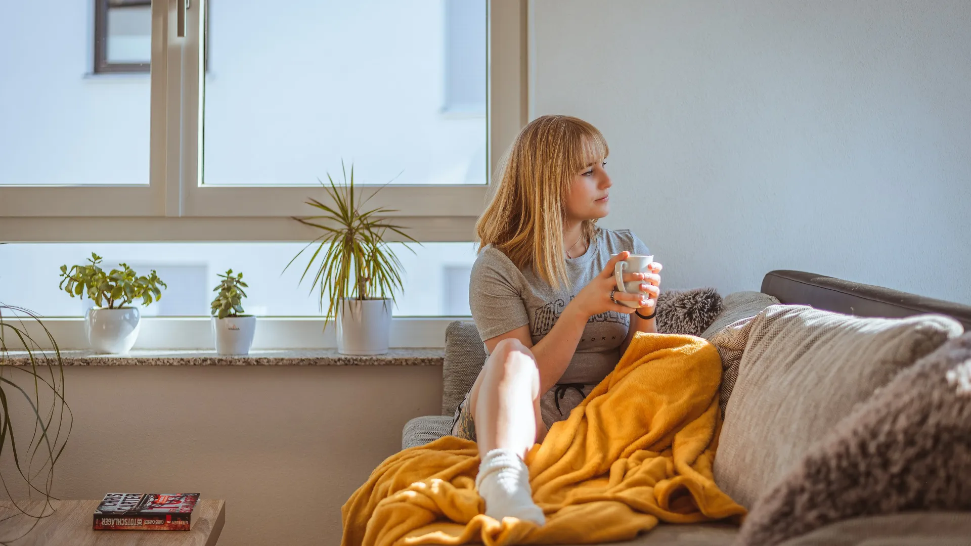 Woman sitting on couch wrapped in yellow blanket, holding mug and looking out window in cozy living room.