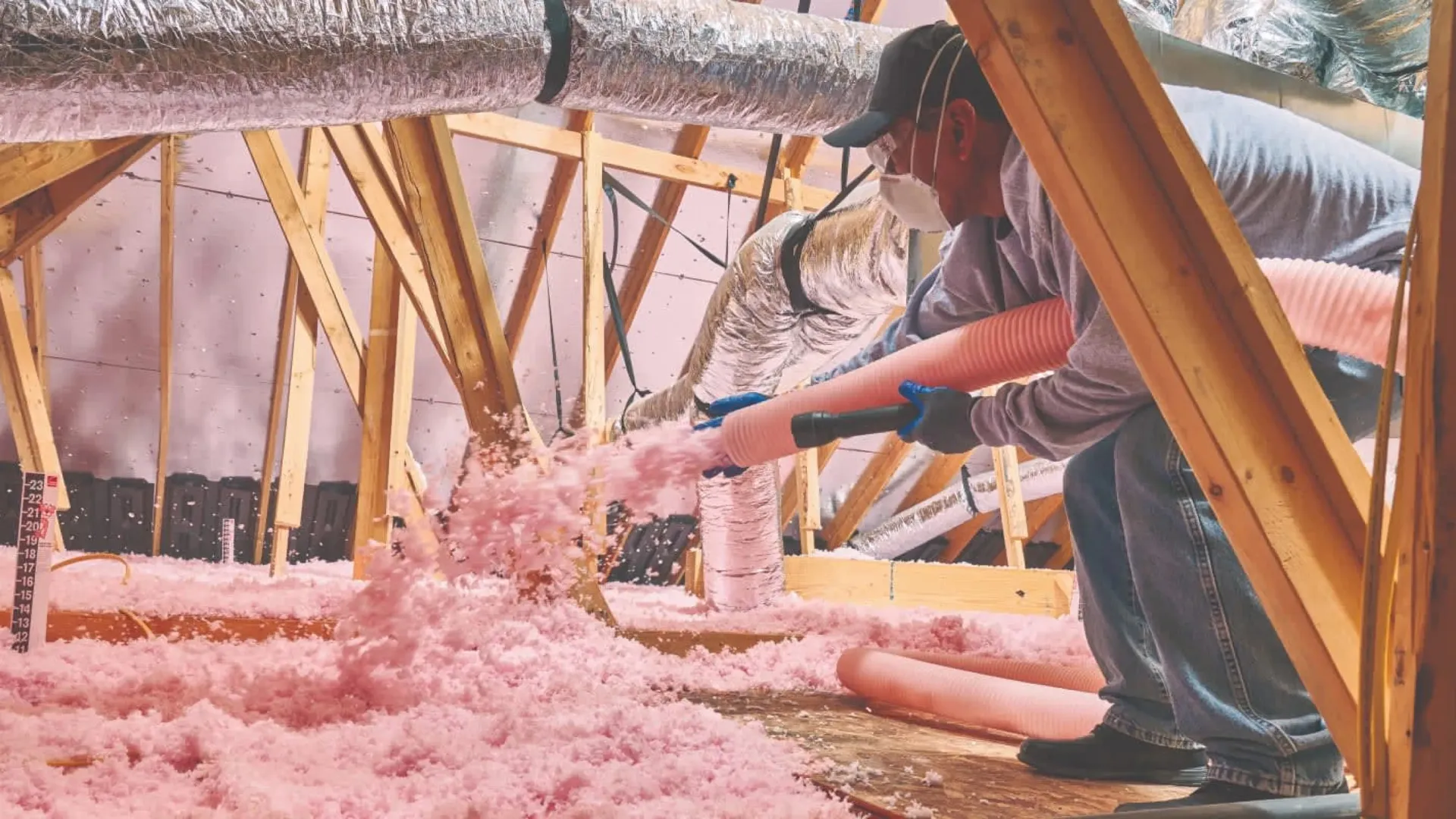 Worker installing pink fiberglass insulation in attic using blower hose and protective mask and gloves