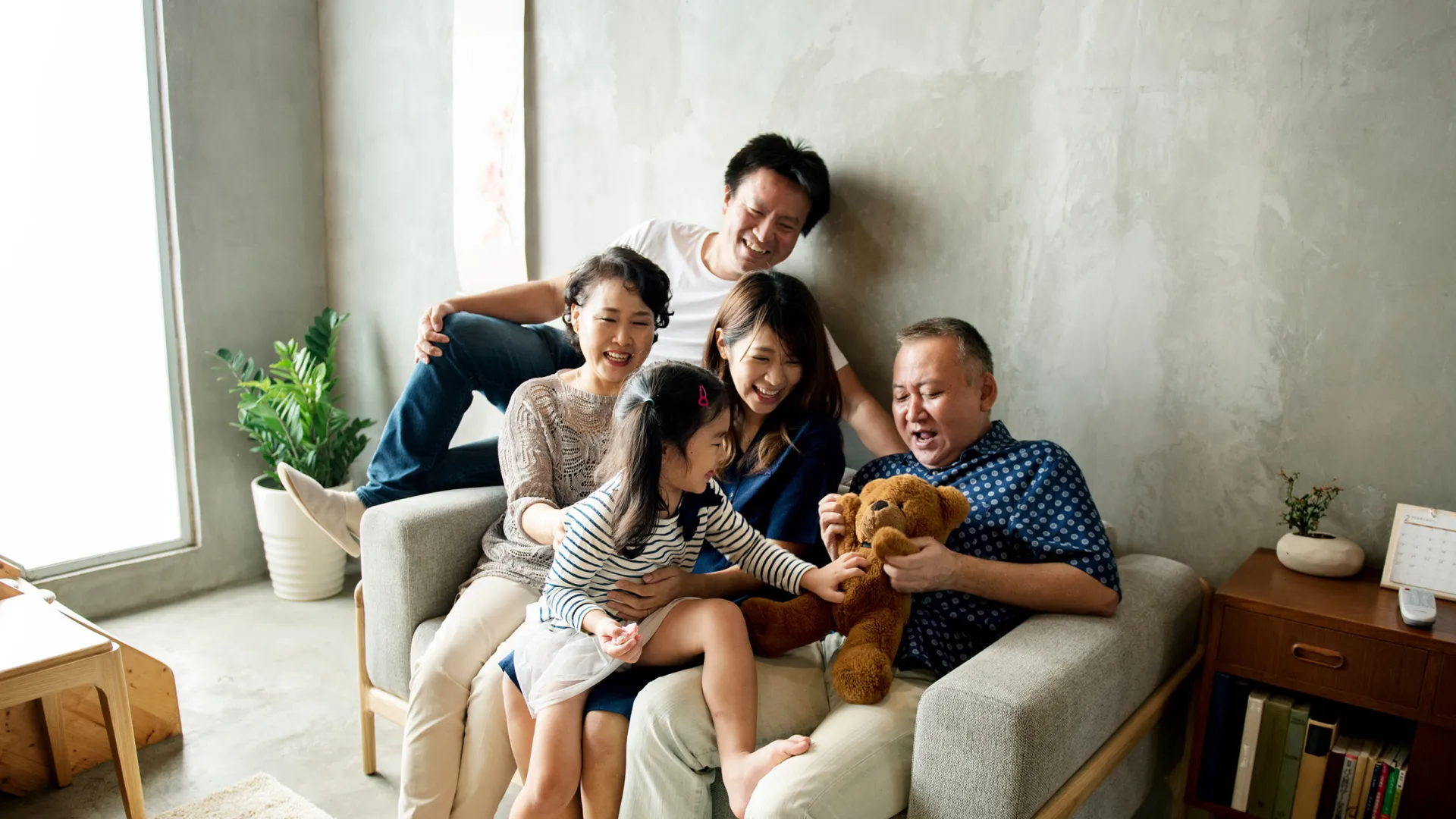 Happy multi-generation Asian family sitting on a couch playing with a brown teddy bear in a bright living room.